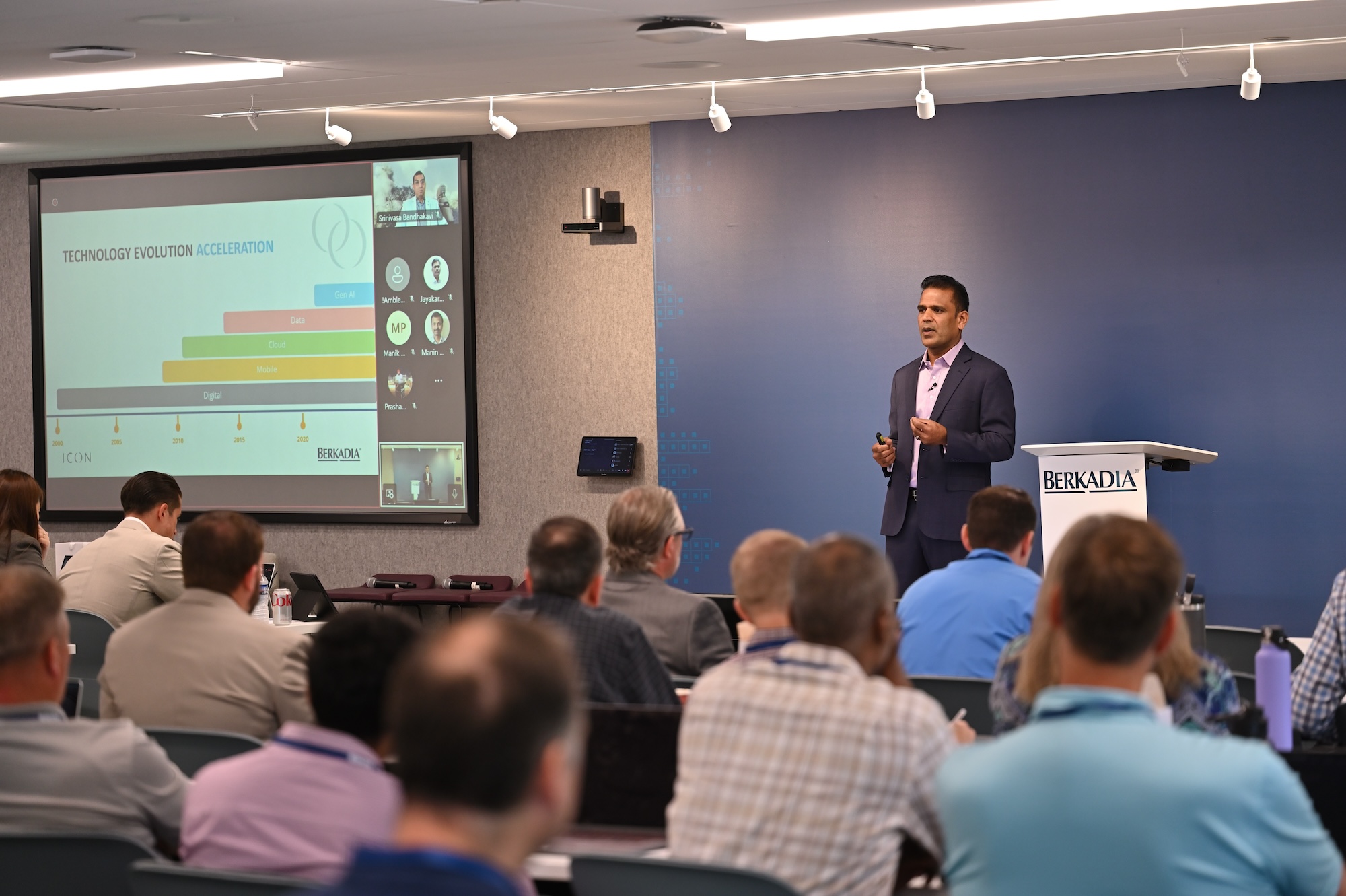 A presenter speaks to an audience in a conference room, standing by a podium and a screen displaying a technology evolution chart.