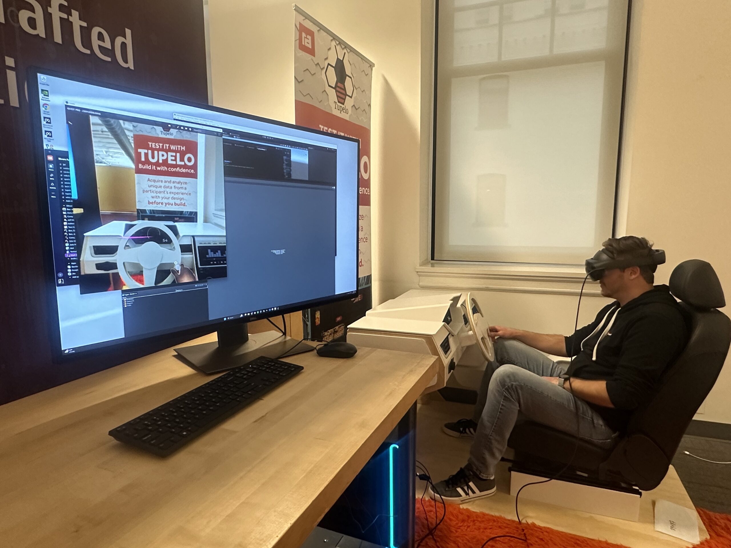 A man wearing a VR headset sits behind a fake steering wheel. A computer next to him displays what he sees of the fake steering wheel through the VR headset.