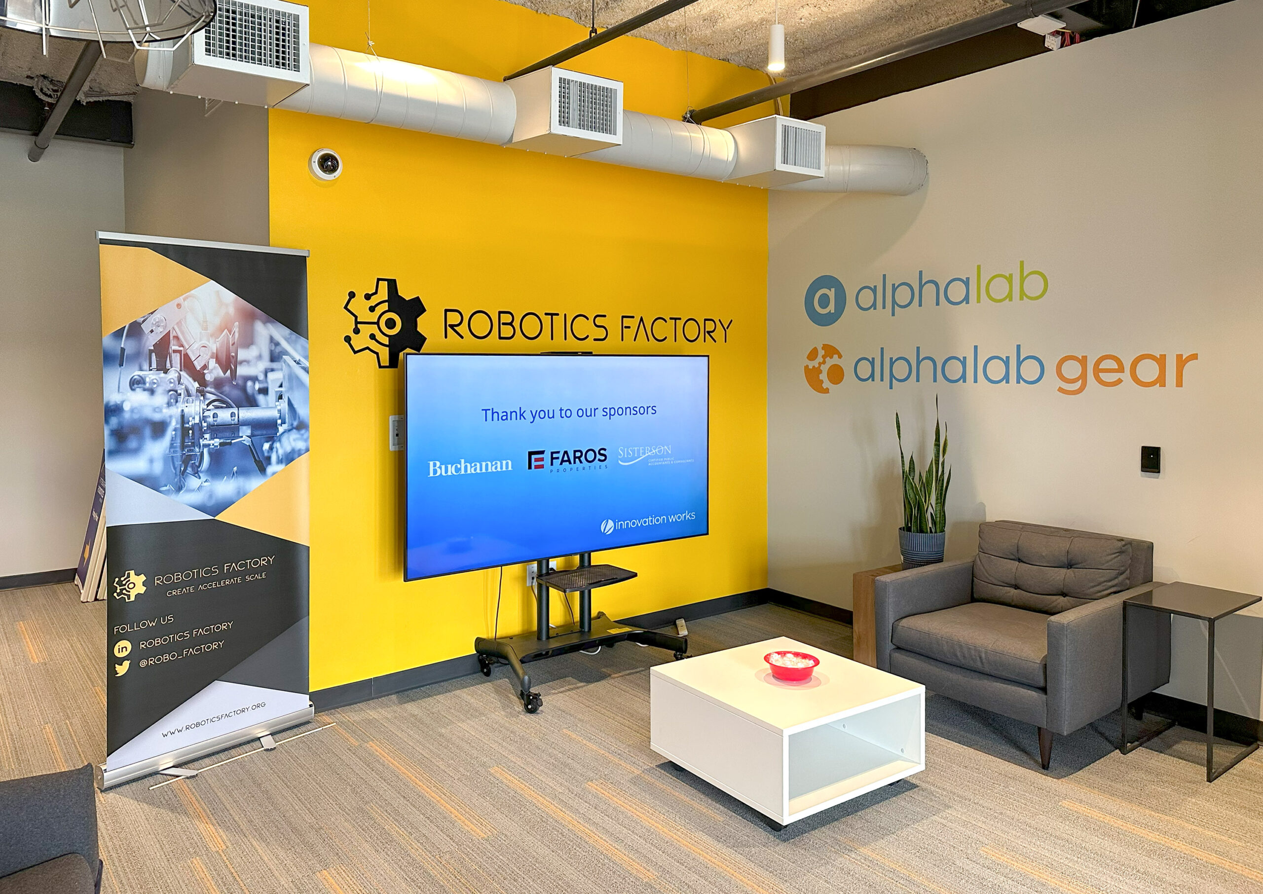 A room with a yellow accent wall, TV displaying sponsor names, a standing banner, and a seating area with a gray chair and white table. Logos for "alphalab" and "alphalab gear" on the wall.