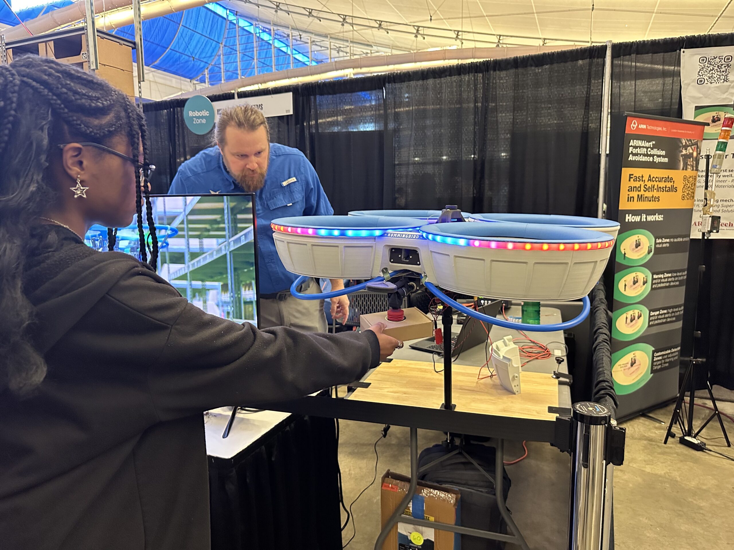 A person interacts with a drone displayed on a table. Another person stands behind the table with informational posters and a screen nearby.