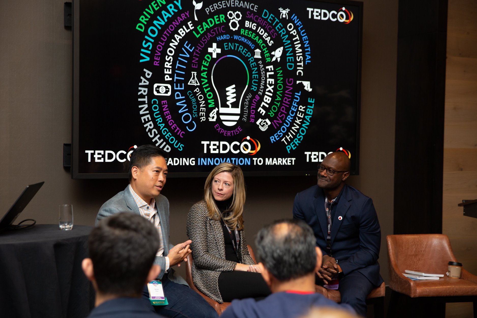 Three people seated, engaging in a panel discussion. A screen behind them displays words like "innovation" and "entrepreneurship" inside a light bulb graphic. Audience members are visible in the foreground.
