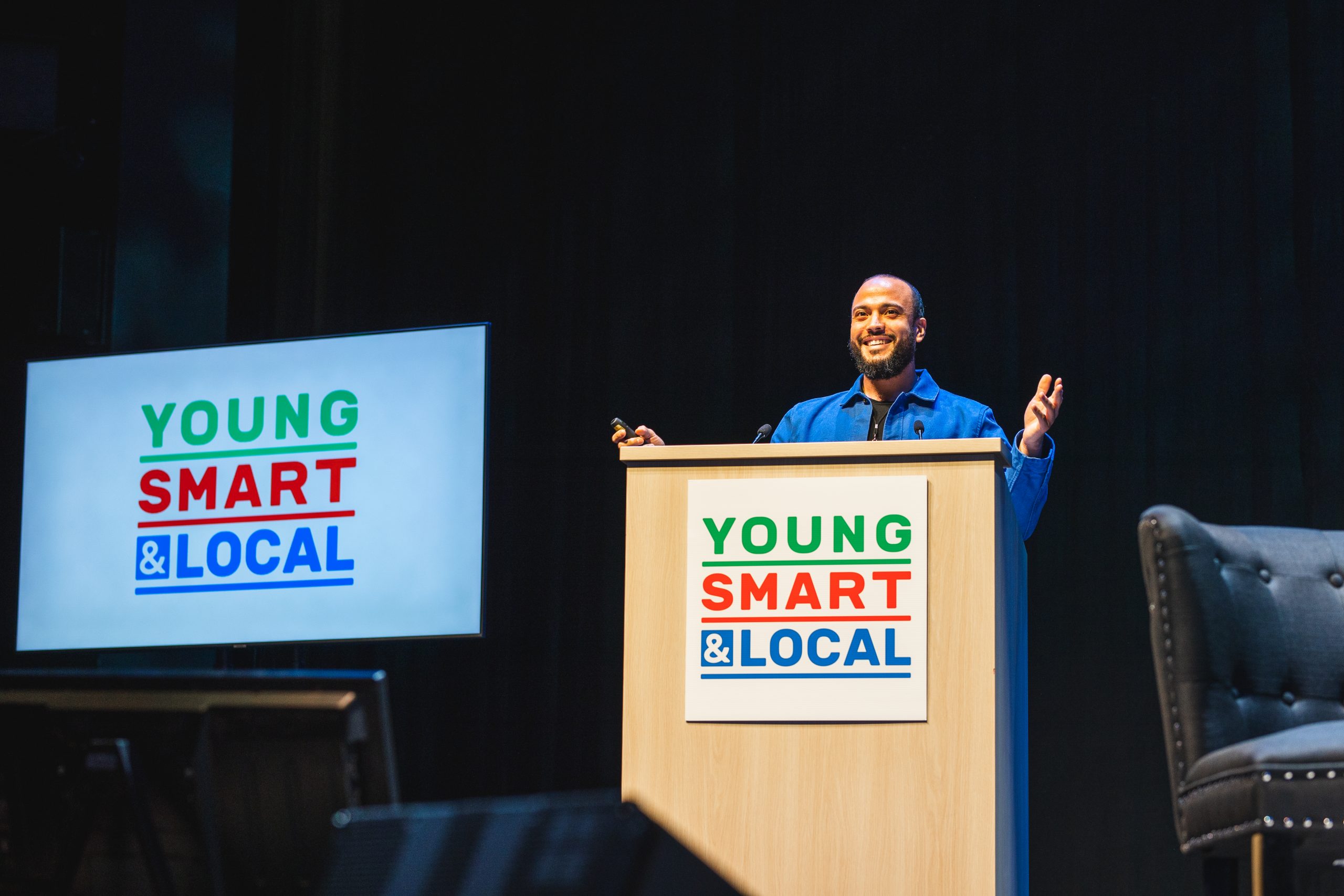 A person stands at a podium labeled "Young Smart & Local," speaking at an event. A screen displays the same text.