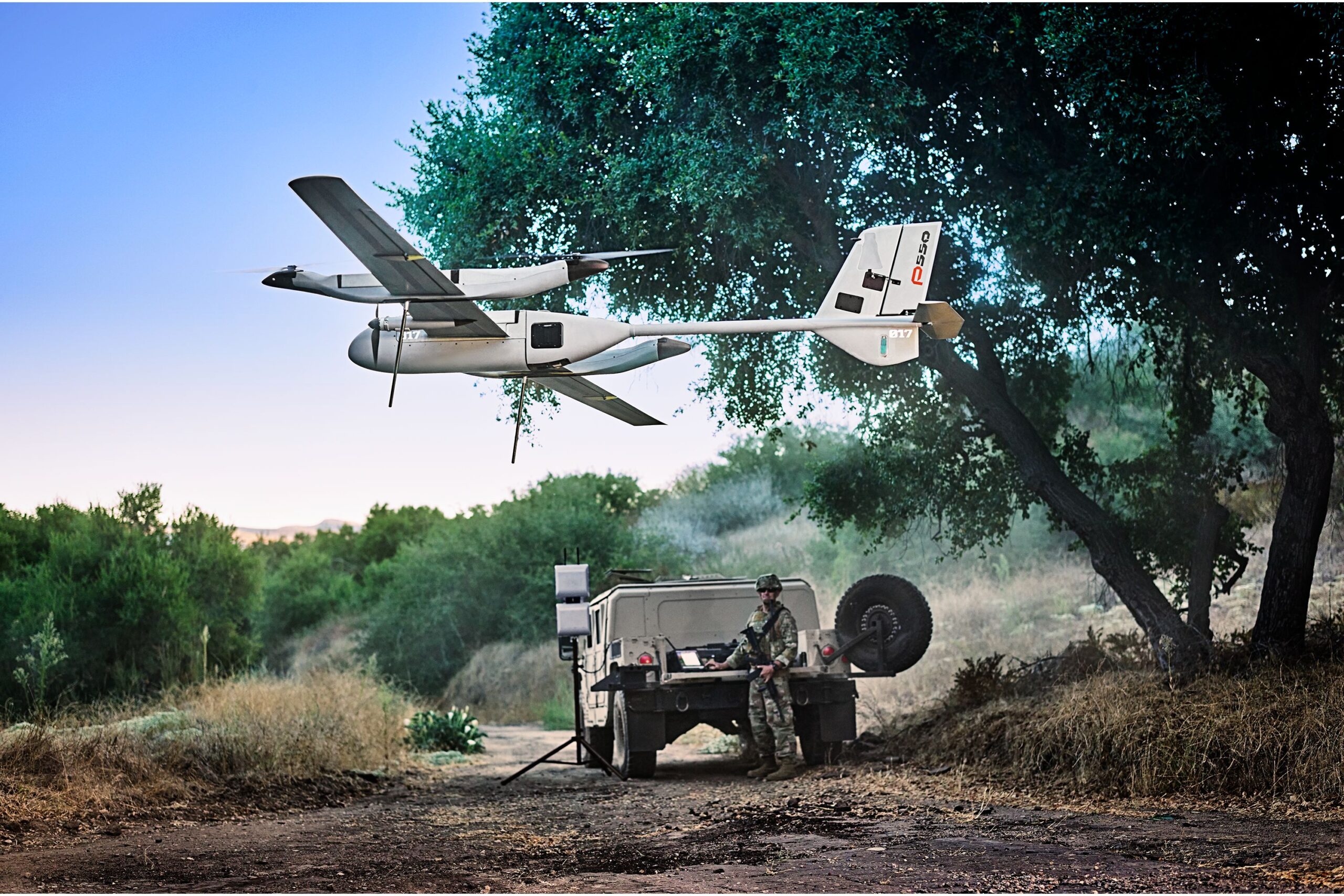 A brown Humvee sits behind an aerial device flying before green trees and brown dirt.