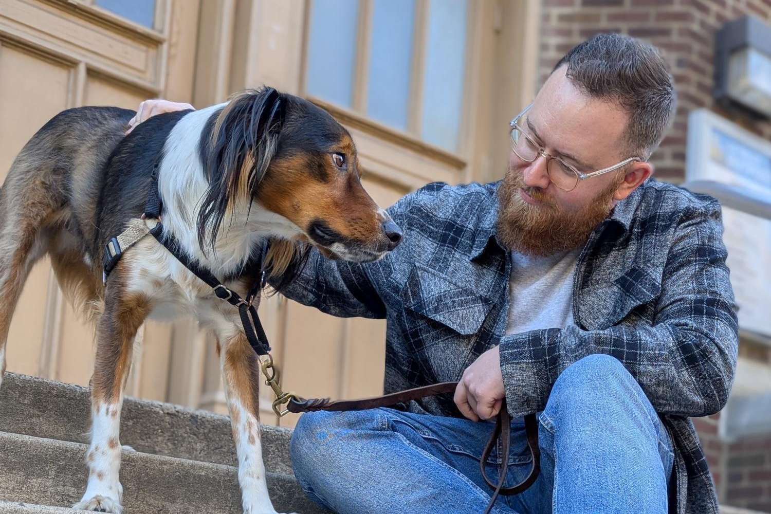A man with a beard pets a dog on a leash while sitting on outdoor steps.