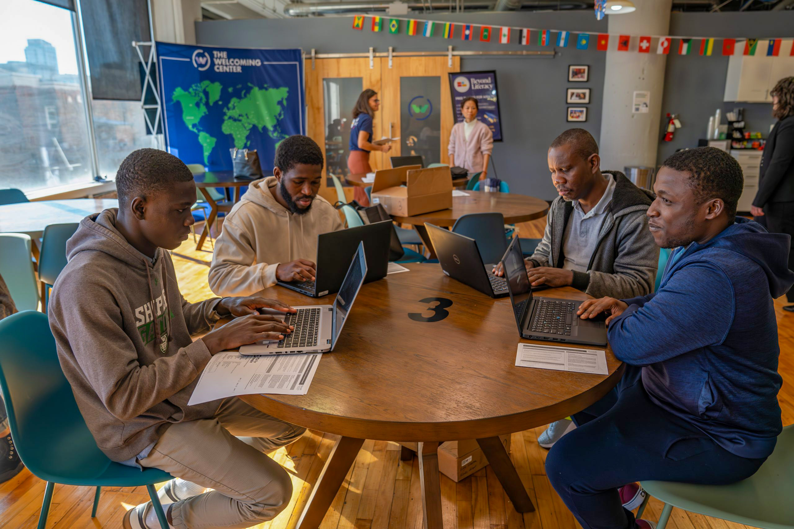 Four people sitting around a round table use laptops in a communal workspace, with others interacting in the background. Flags and informational posters are displayed on the walls.