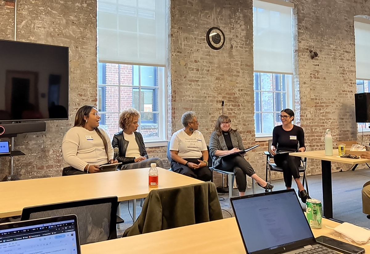 A panel of five people sitting in front of a brick wall with windows, engaged in a discussion. Tables with laptops and drinks are in the foreground.