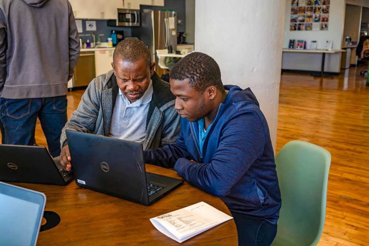 Two people, an older Black man and young Black man, are seated at a table working on laptops. A notebook lies open on the table. They are in an office setting.