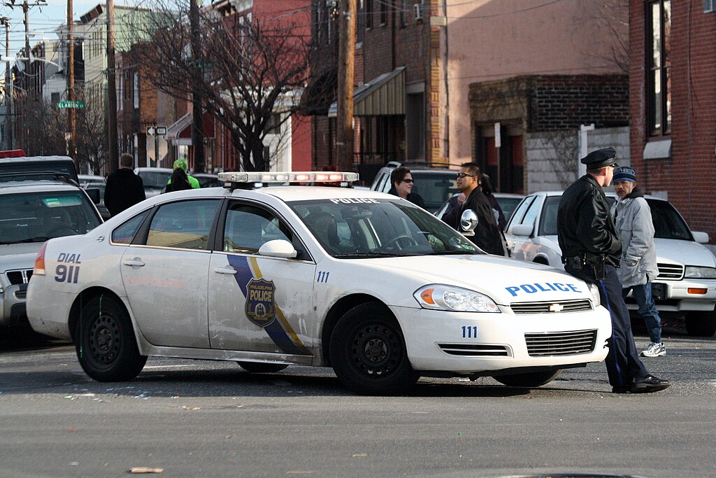 A police car is parked on a city street with several officers and people nearby. Buildings line the background.