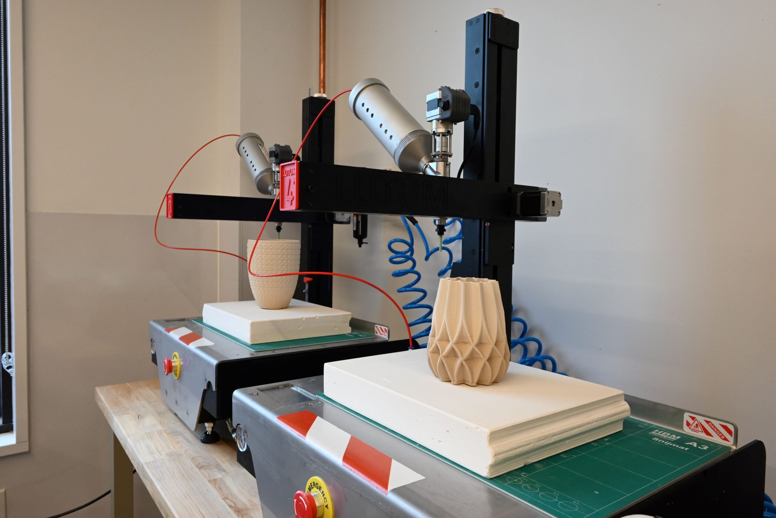 Two 3D printers creating vases on a workbench, with red wires and geometric designs on the vases.