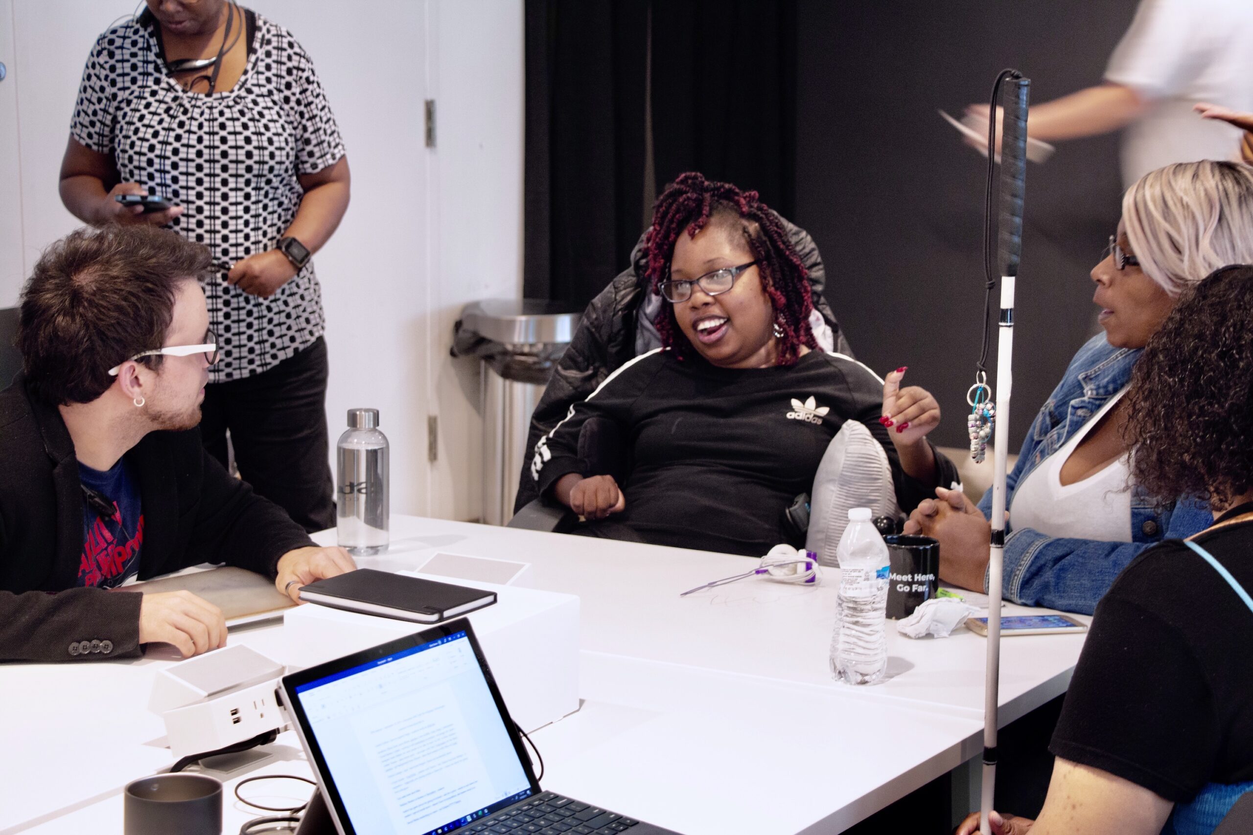 People stand and sit around an off-white conference table with laptops, notebooks and other materials on it