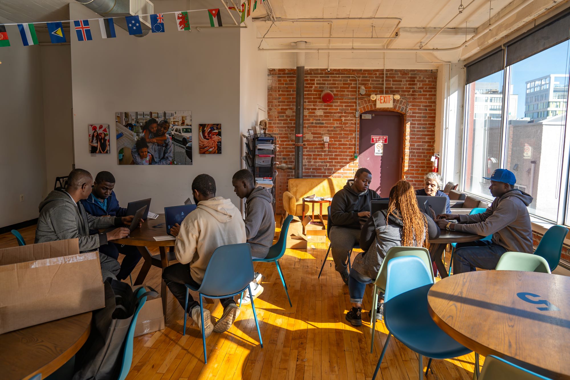 A group of people sits at tables in a room, using laptops. Sunlight streams through large windows, and various flags hang from the ceiling.