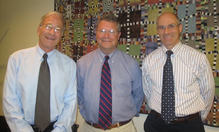 Three men in dress shirts and ties stand in front of a colorful, geometric-patterned quilt.
