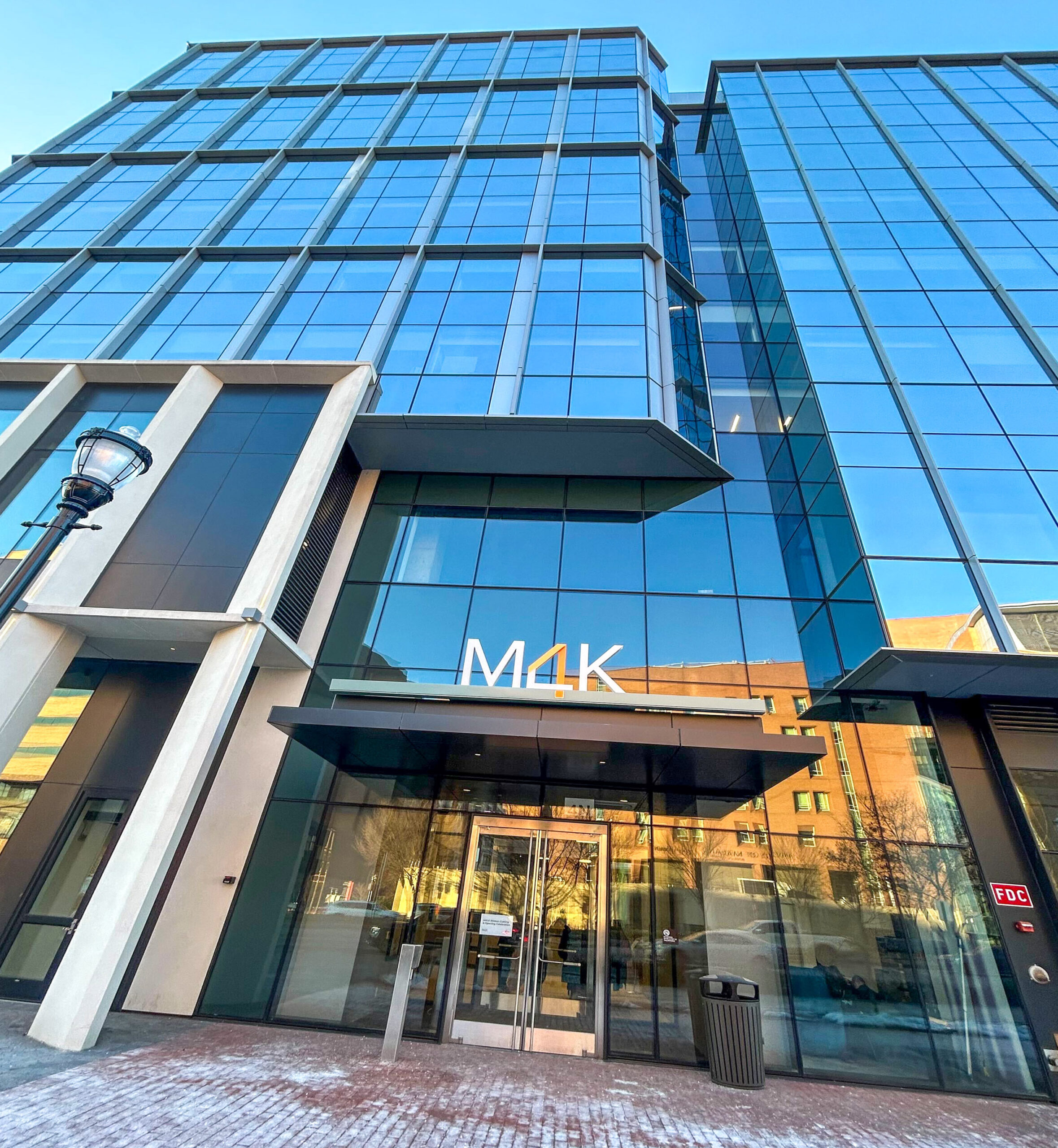 A glass-fronted building looms over a red brick sidewalk with white snow. "M4K" is spelled in white and orange over the awning.