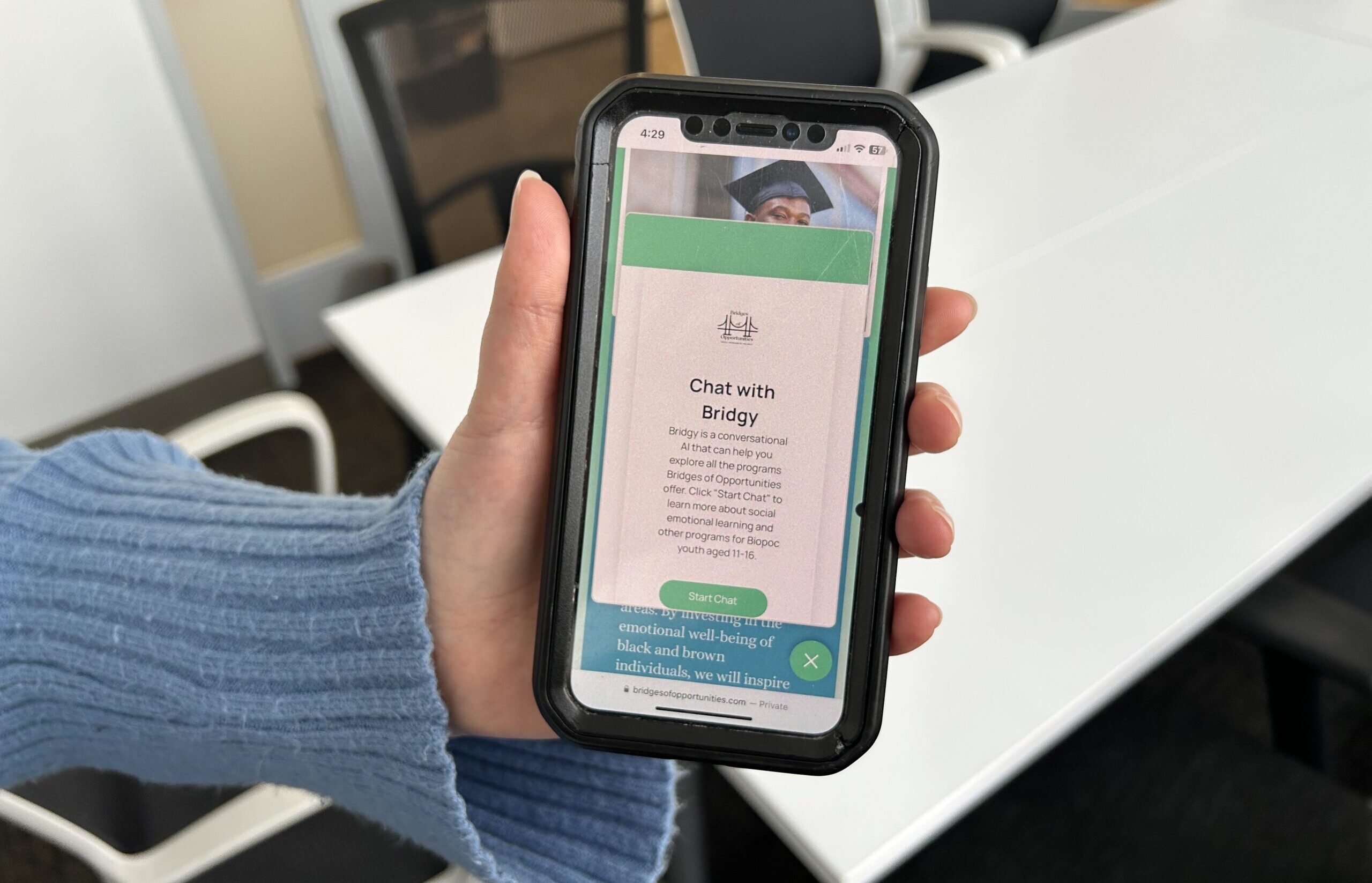 Hand holding a smartphone displaying a chat application named "Bridgy" with text about emotional well-being. The background shows an office setting with a white table and chairs.