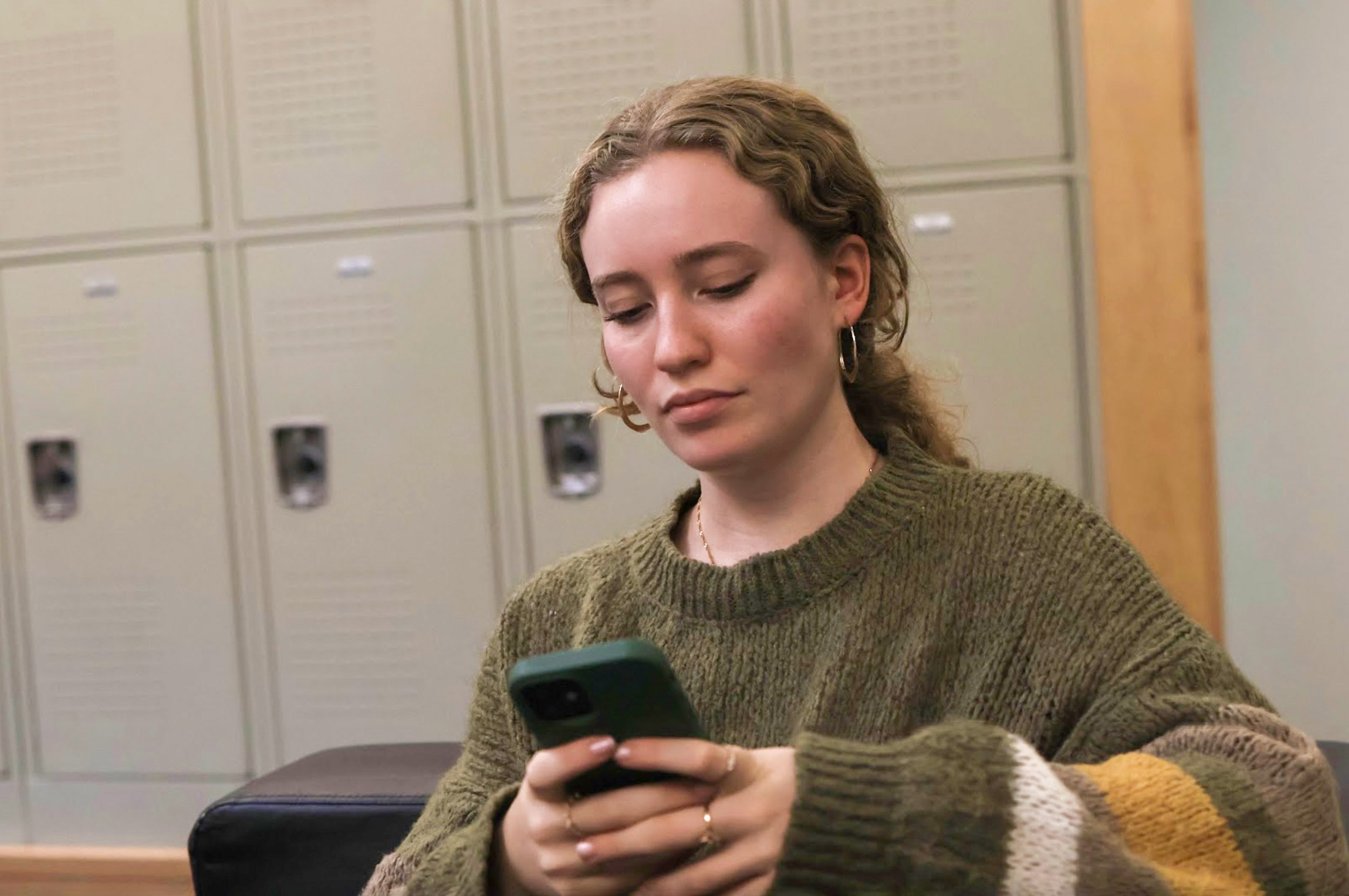 Teenager wearing a green sweater, looking at a smartphone in front of a row of lockers.