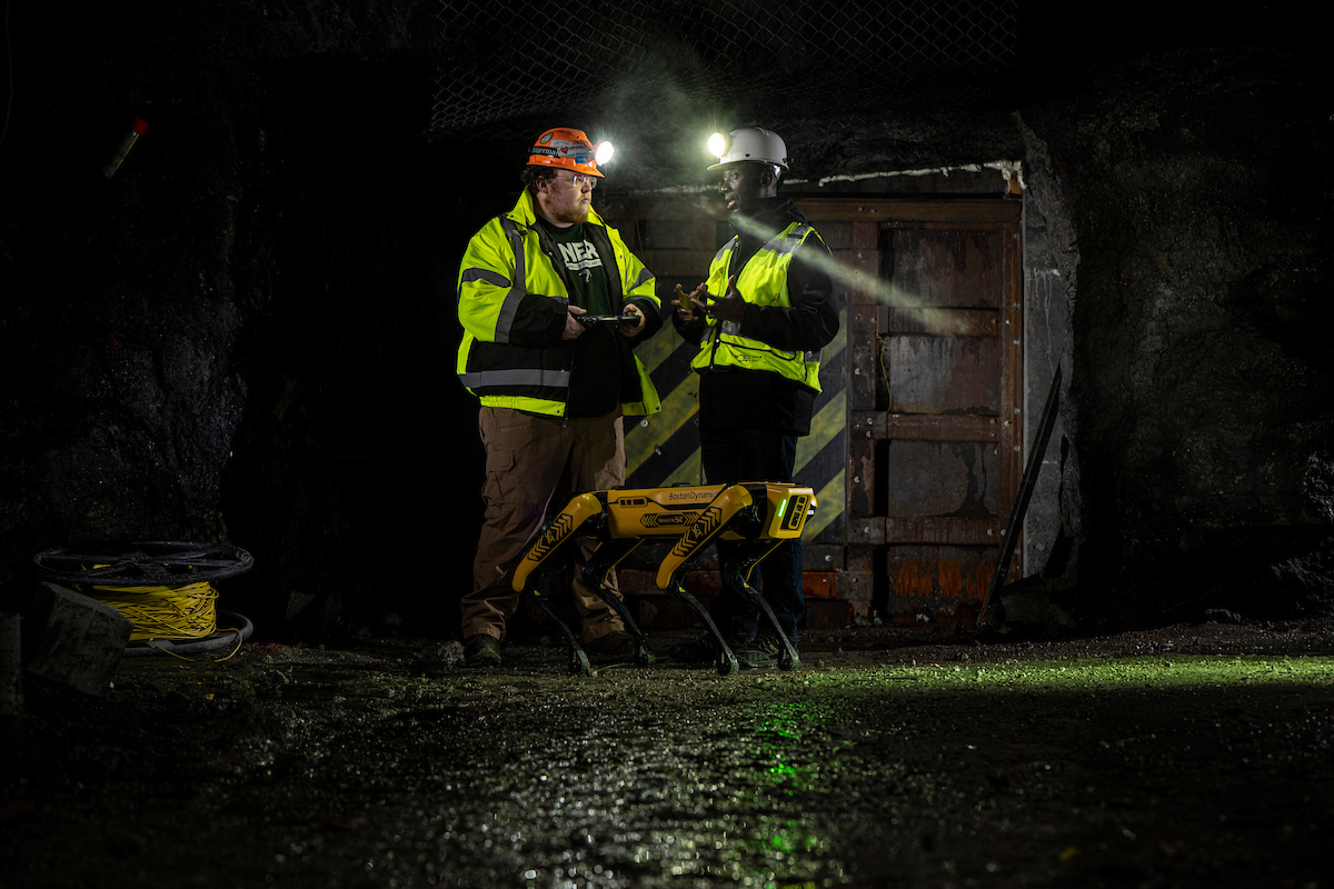 Two workers in safety gear converse with a robotic dog in a dimly lit underground environment, with helmets and safety jackets visible.