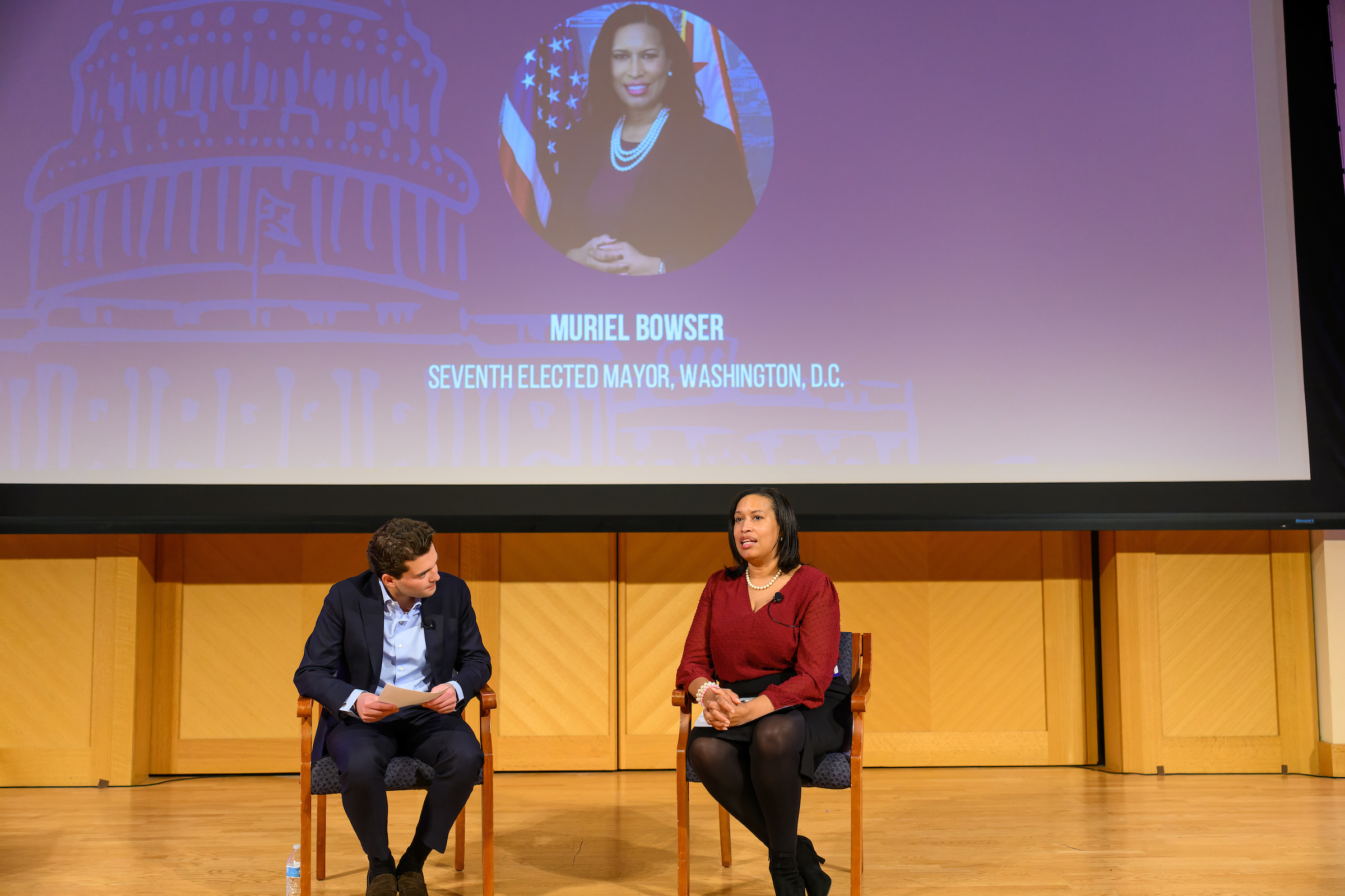 Two people sitting on a stage, one speaking into a microphone. A large screen behind them displays a portrait with the name "Muriel Bowser" and her title as the seventh elected Mayor of Washington, D.C.