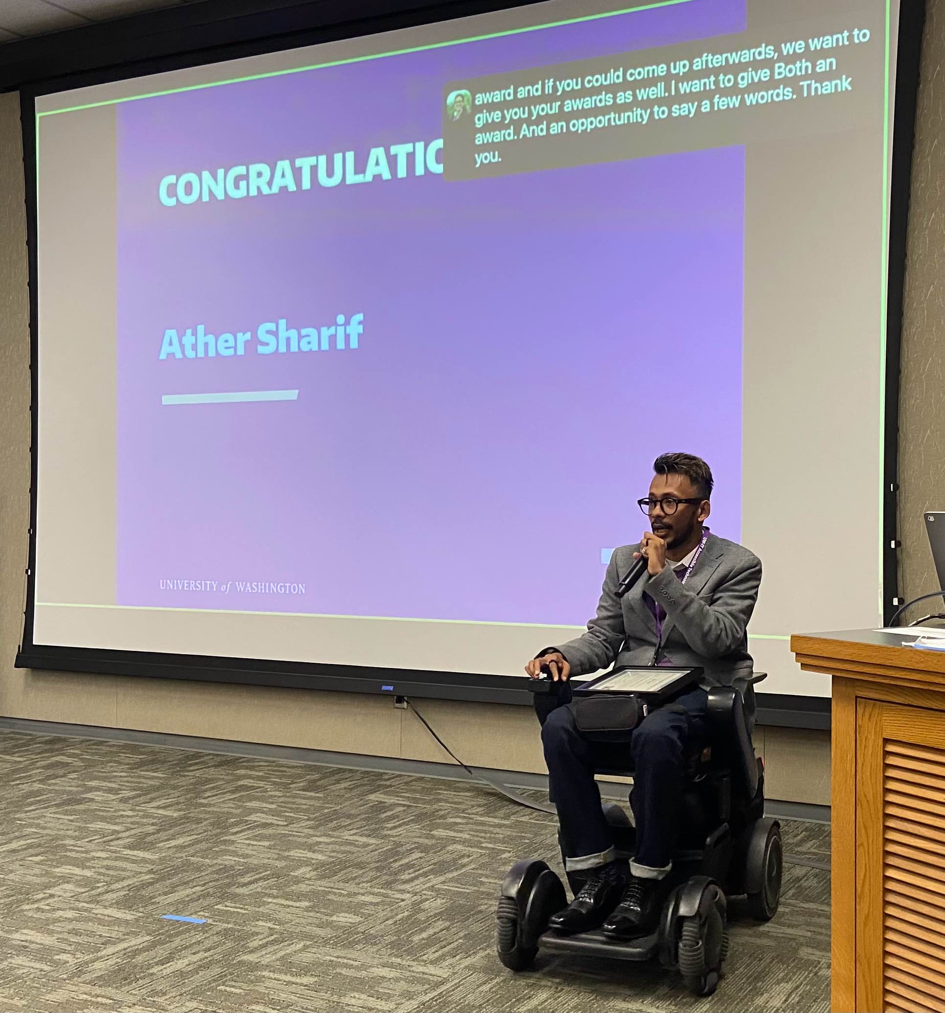 A South Asian man in a black wheelchair speaks in front of a screen with a blue background and white text