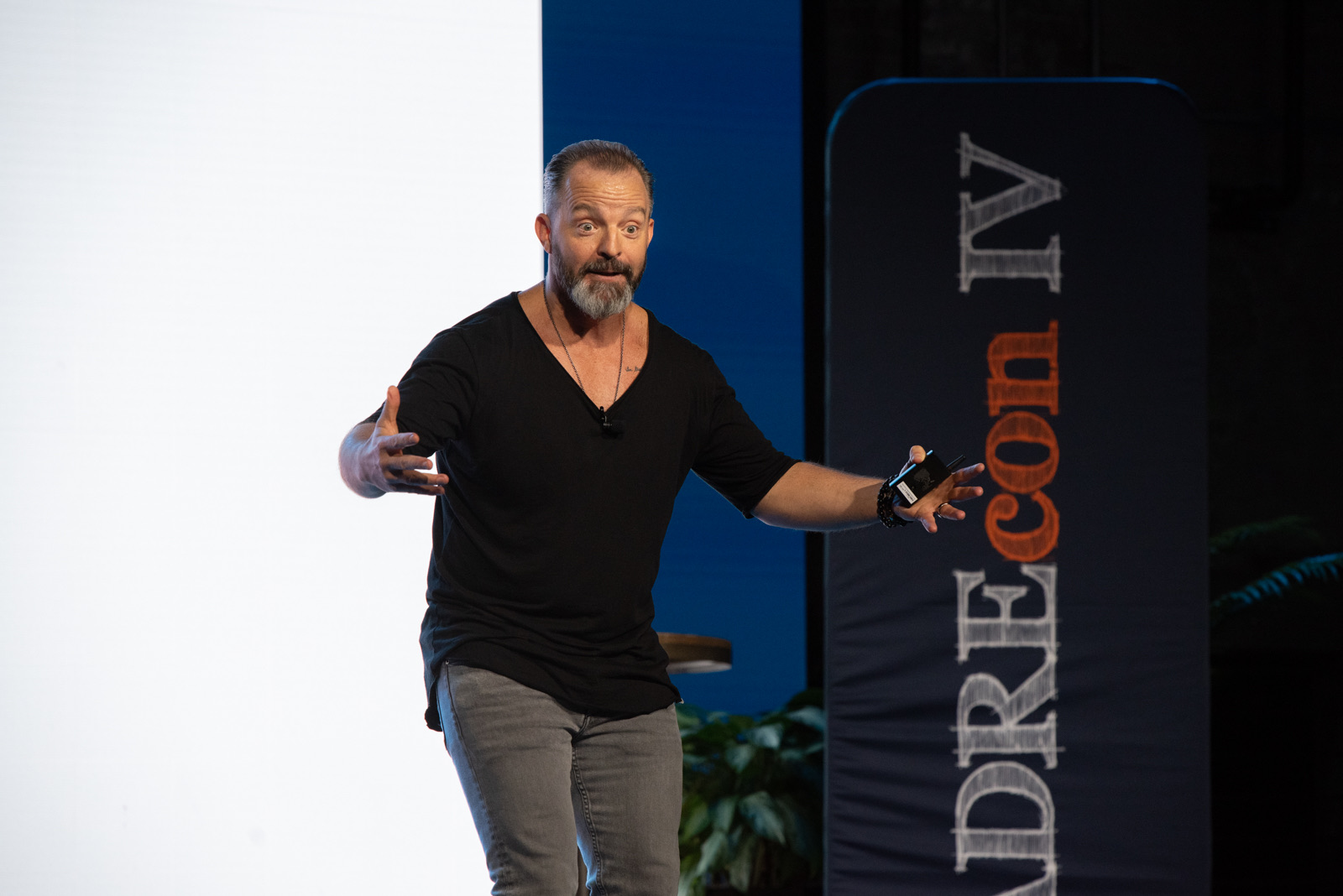 A white man speaks with arms outstretched while holding a black presentation controller and wearing a black tshirt and grey jeans