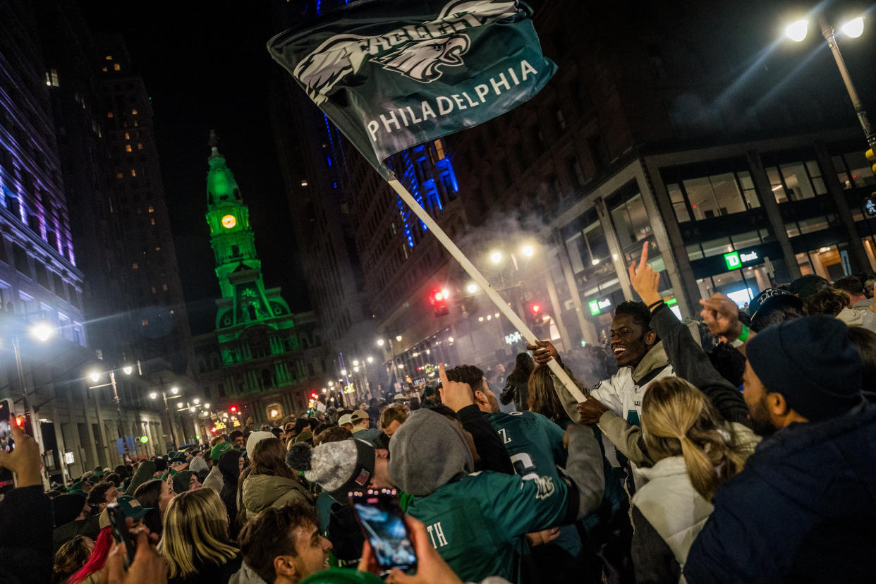A large crowd celebrates on a city street at night, with a person waving a Philadelphia Eagles flag. A building is lit up in green in the background.