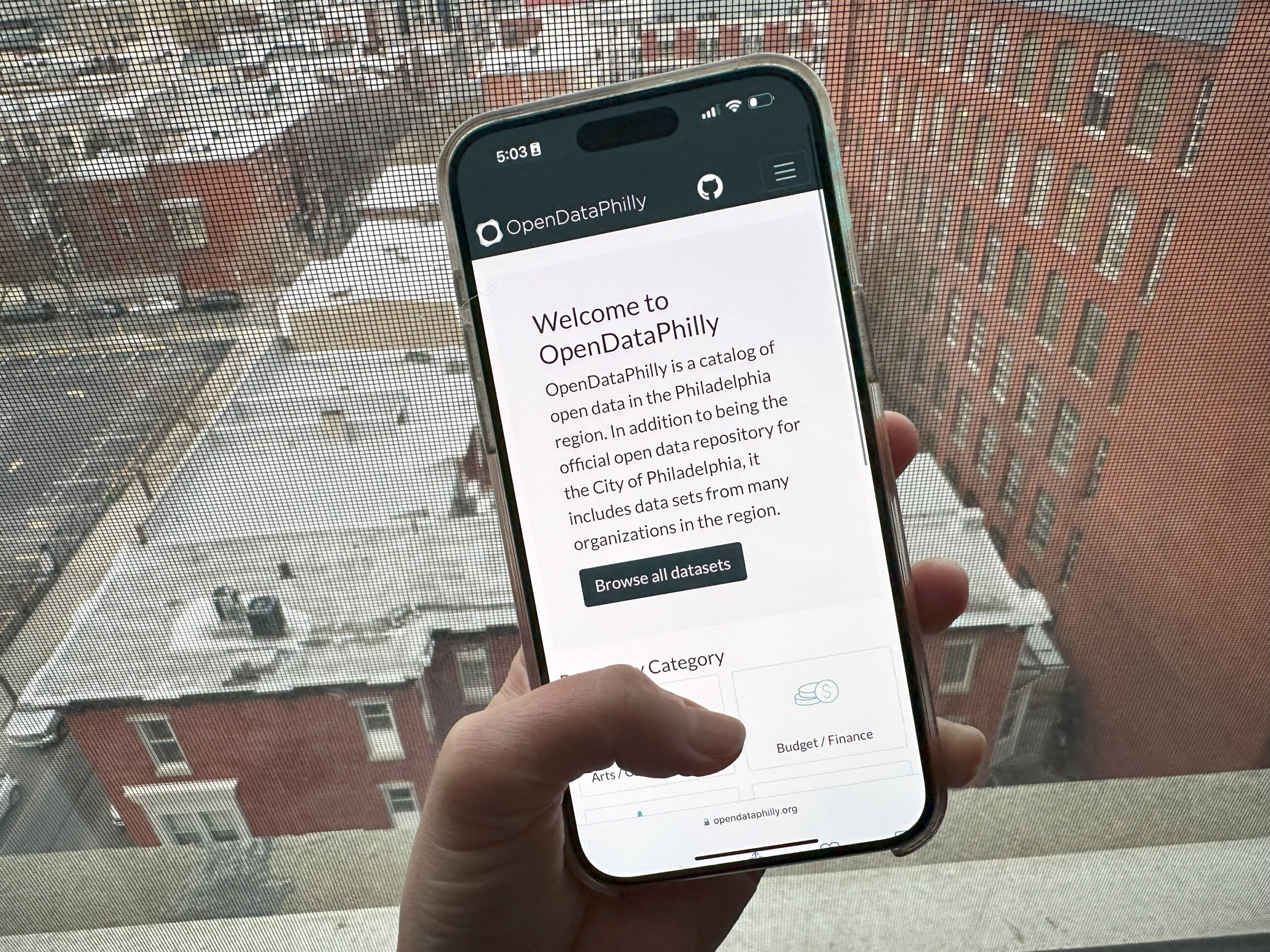 A hand holds a smartphone displaying the OpenDataPhilly website. City buildings can be seen through the window in the background.