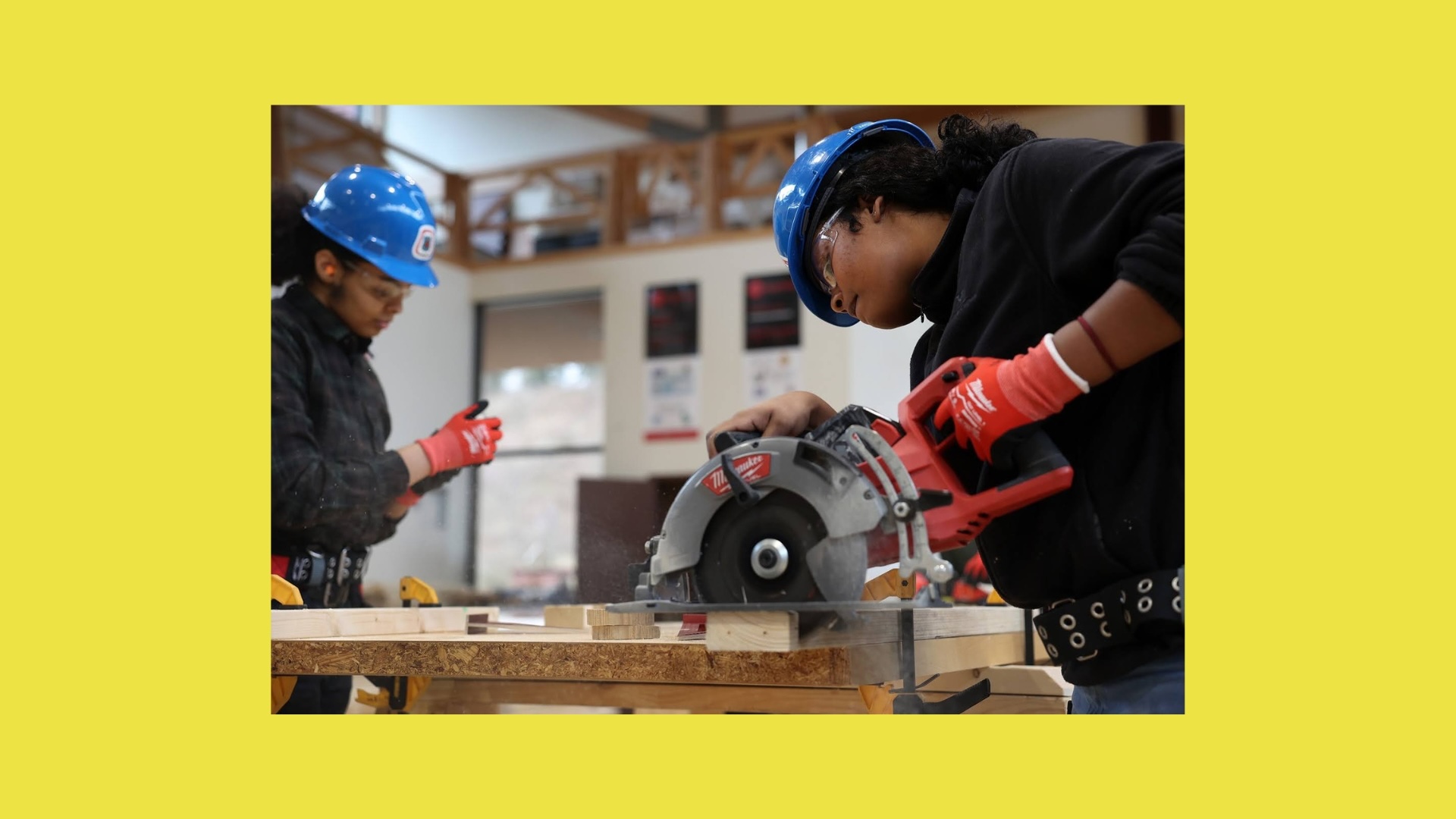 Two people wearing blue helmets and gloves work in a workshop. One uses a circular saw on wooden planks; the other checks a smartphone.
