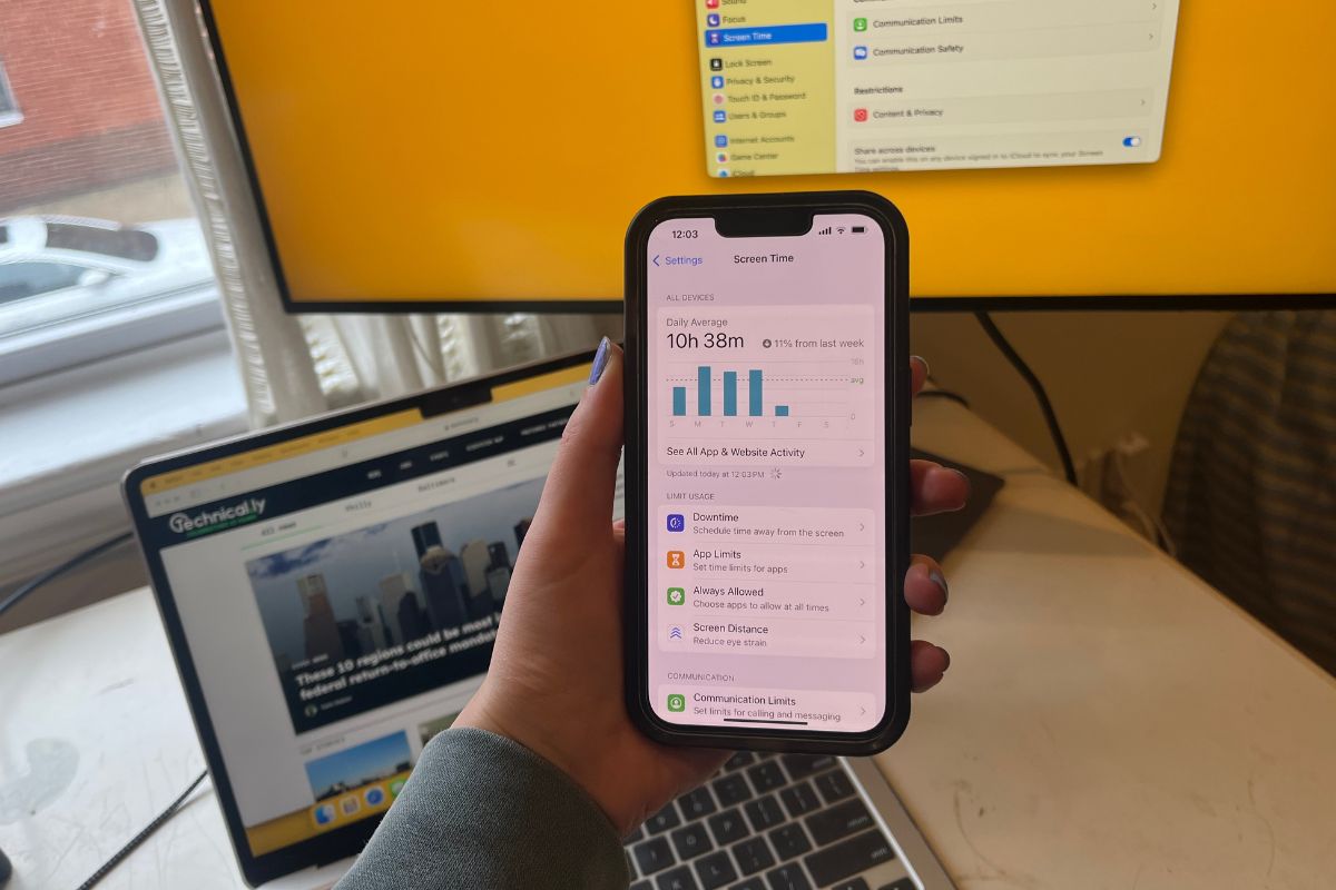 A person holds a smartphone displaying screen time statistics. A laptop is open on a website, while a monitor shows a settings menu.