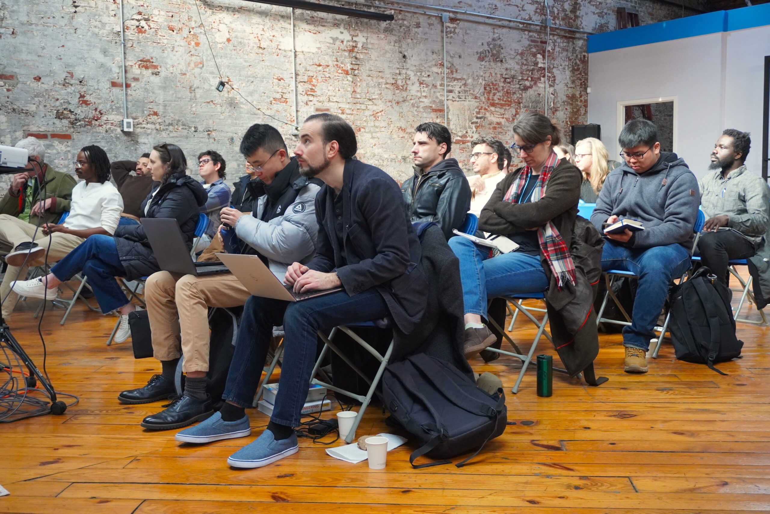 People seated in a room, some with laptops and notebooks, attentively listening to a presentation. The space has wooden floors and exposed brick walls.
