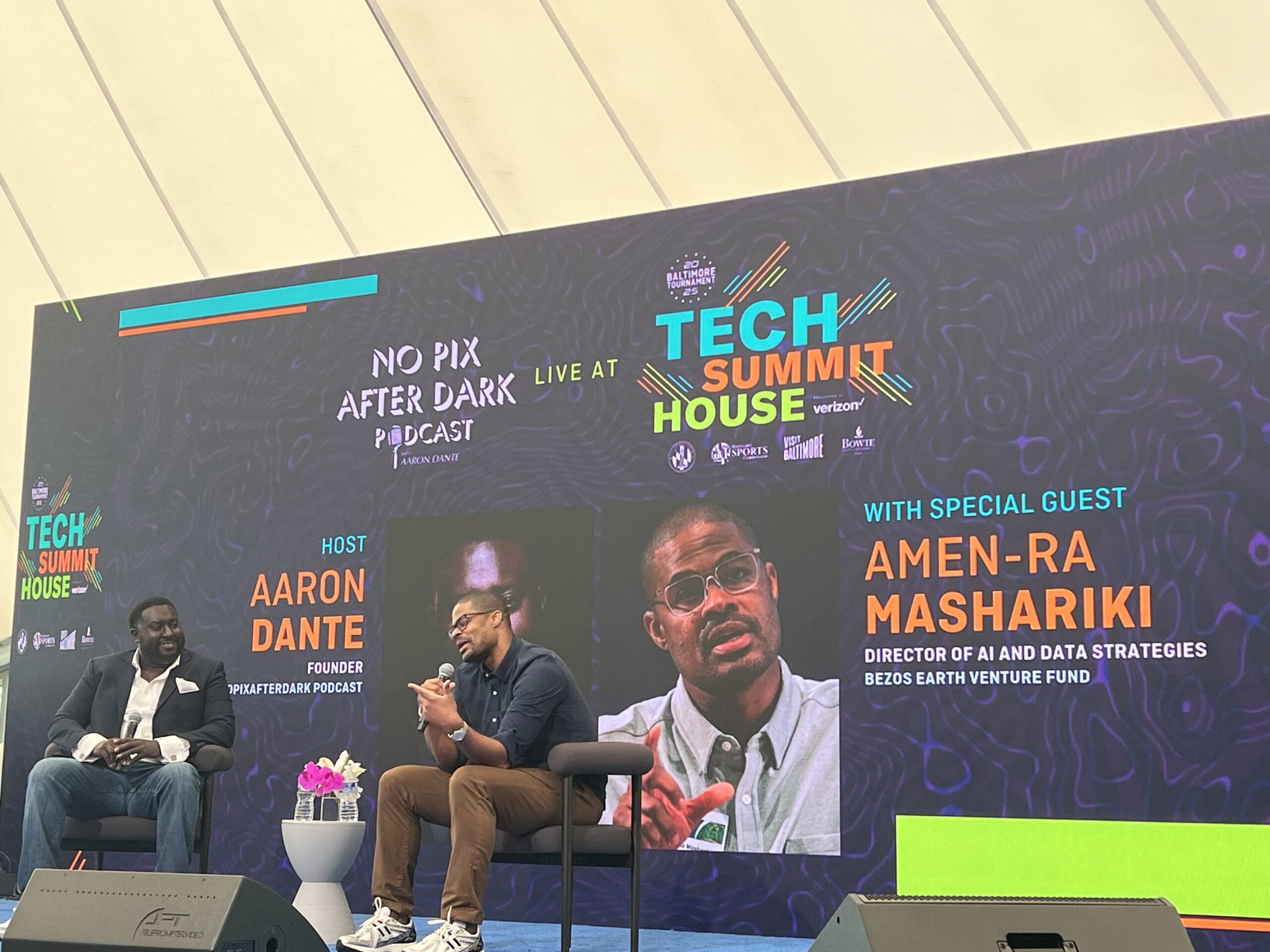 Two men seated on stage engaging in a discussion at Tech Summit House, with a screen displaying their names and roles. Event branding and signage are visible around them.