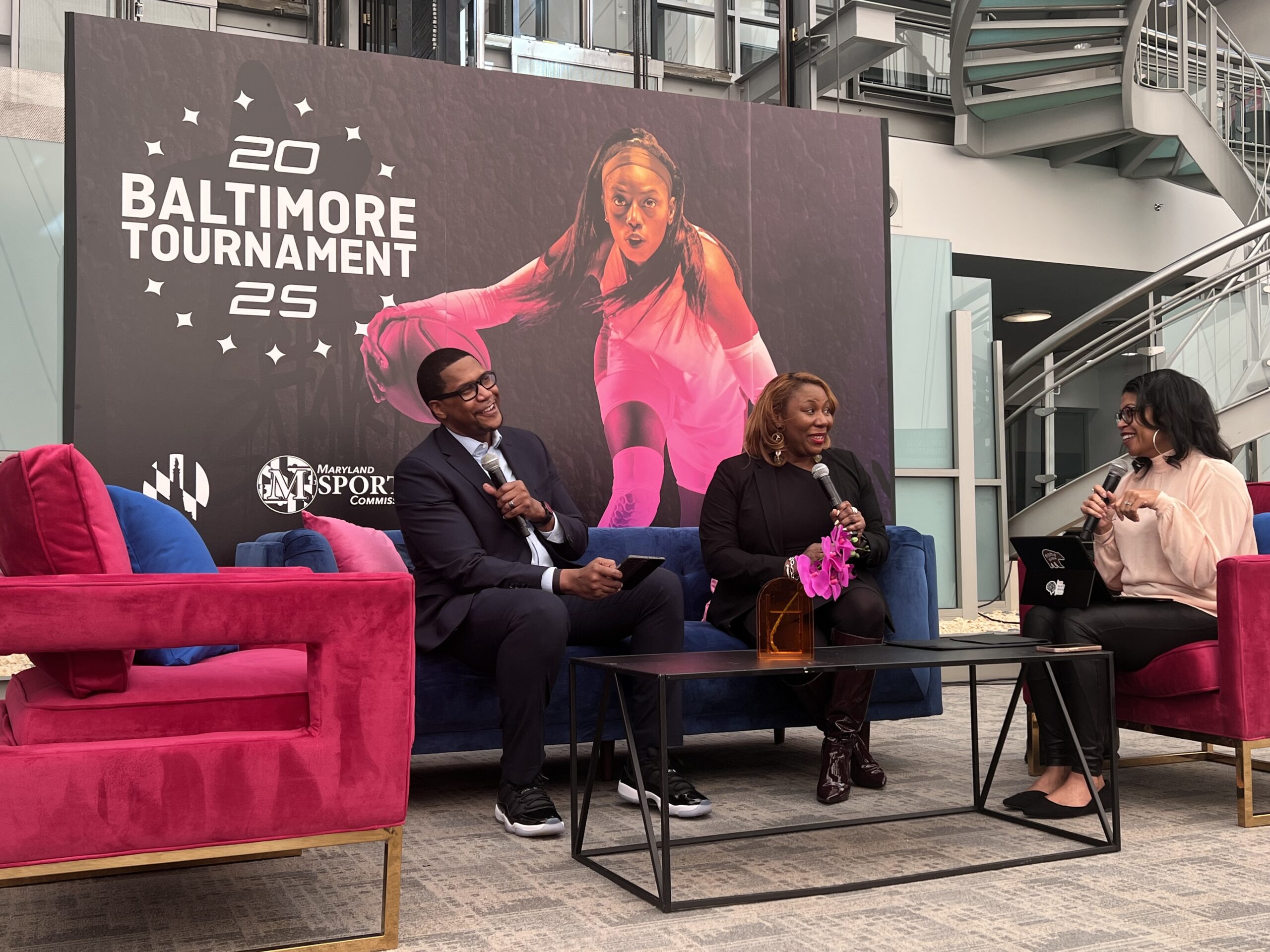 Three people sit in a modern conference area. A man on the left and two women on the right are talking. A poster of a female athlete in action is in the background.