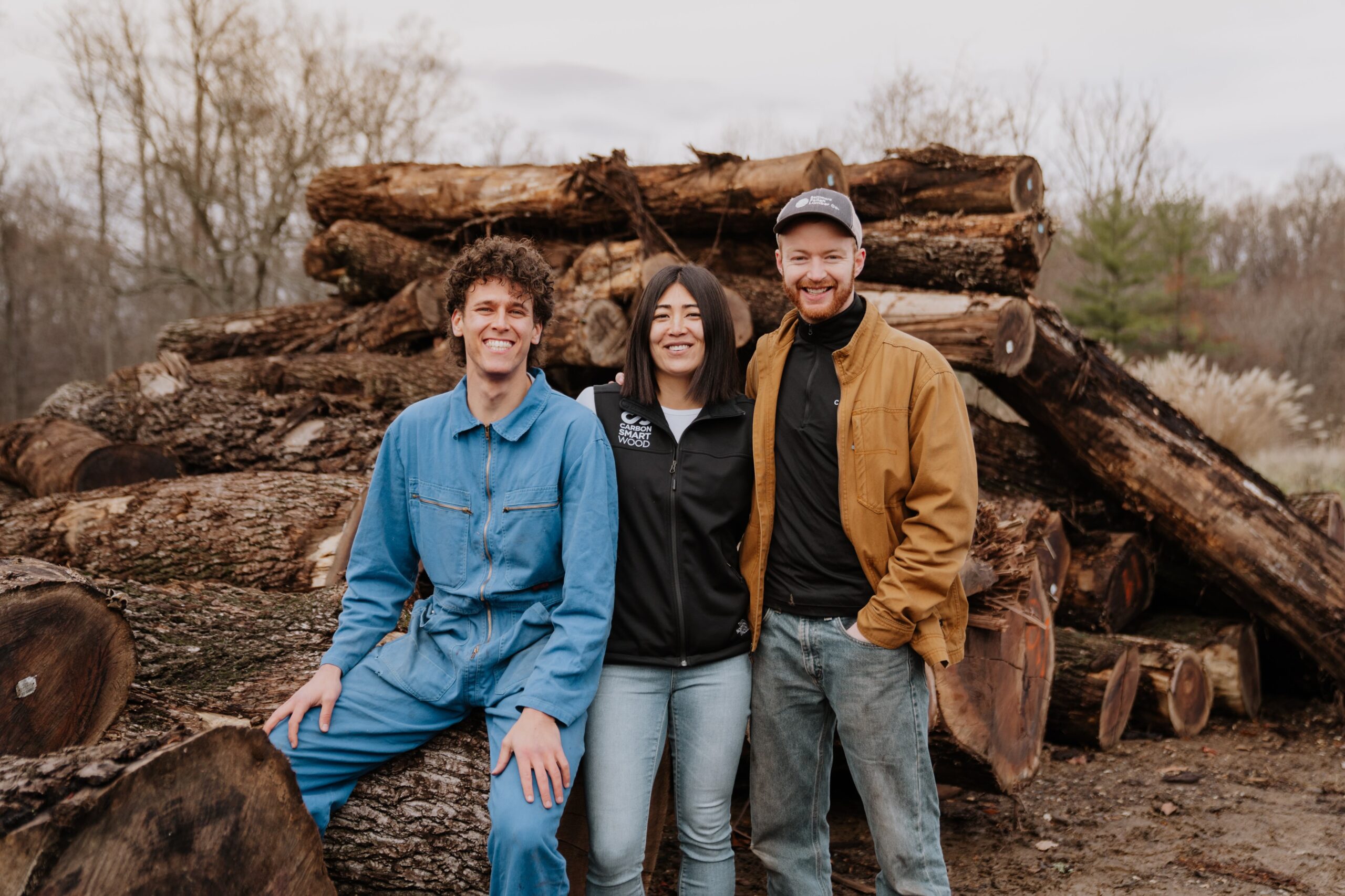 Three people smiling in front of a large pile of logs. One is seated, and two are standing. Outdoors with a cloudy sky.