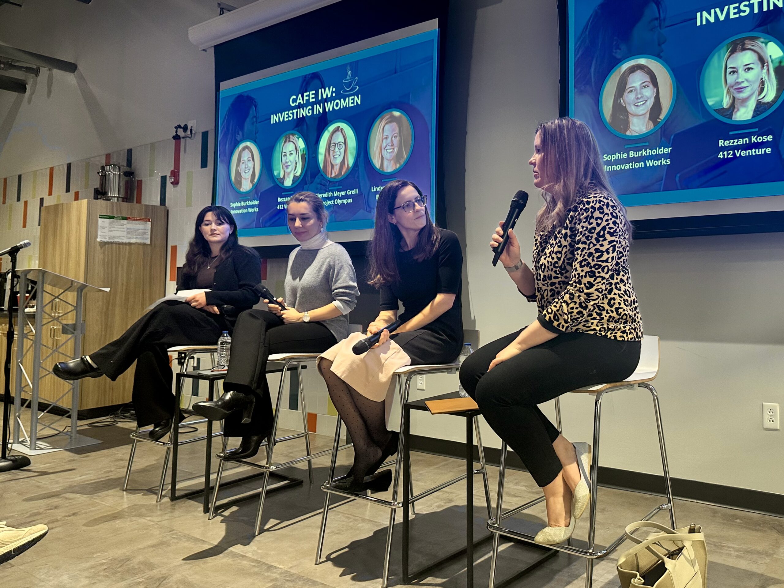 Four women sit on chairs, holding microphones, in front of two screens displaying "Cafe IW: Investing in Women." The woman on the far right is speaking while the others listen attentively.