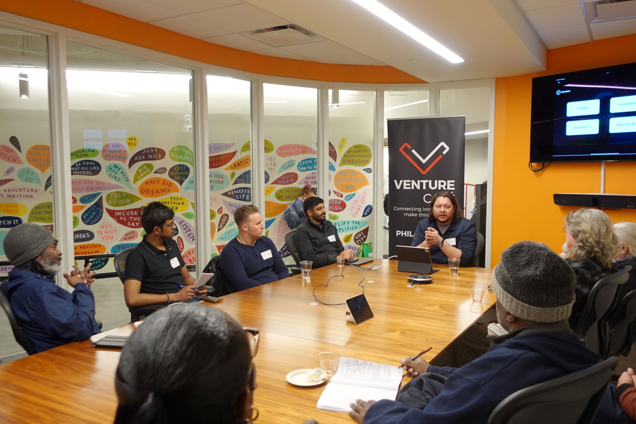 People seated around a conference table in a meeting room with colorful wall art. A speaker addresses the group next to a banner reading "Venture Cafe Philadelphia.