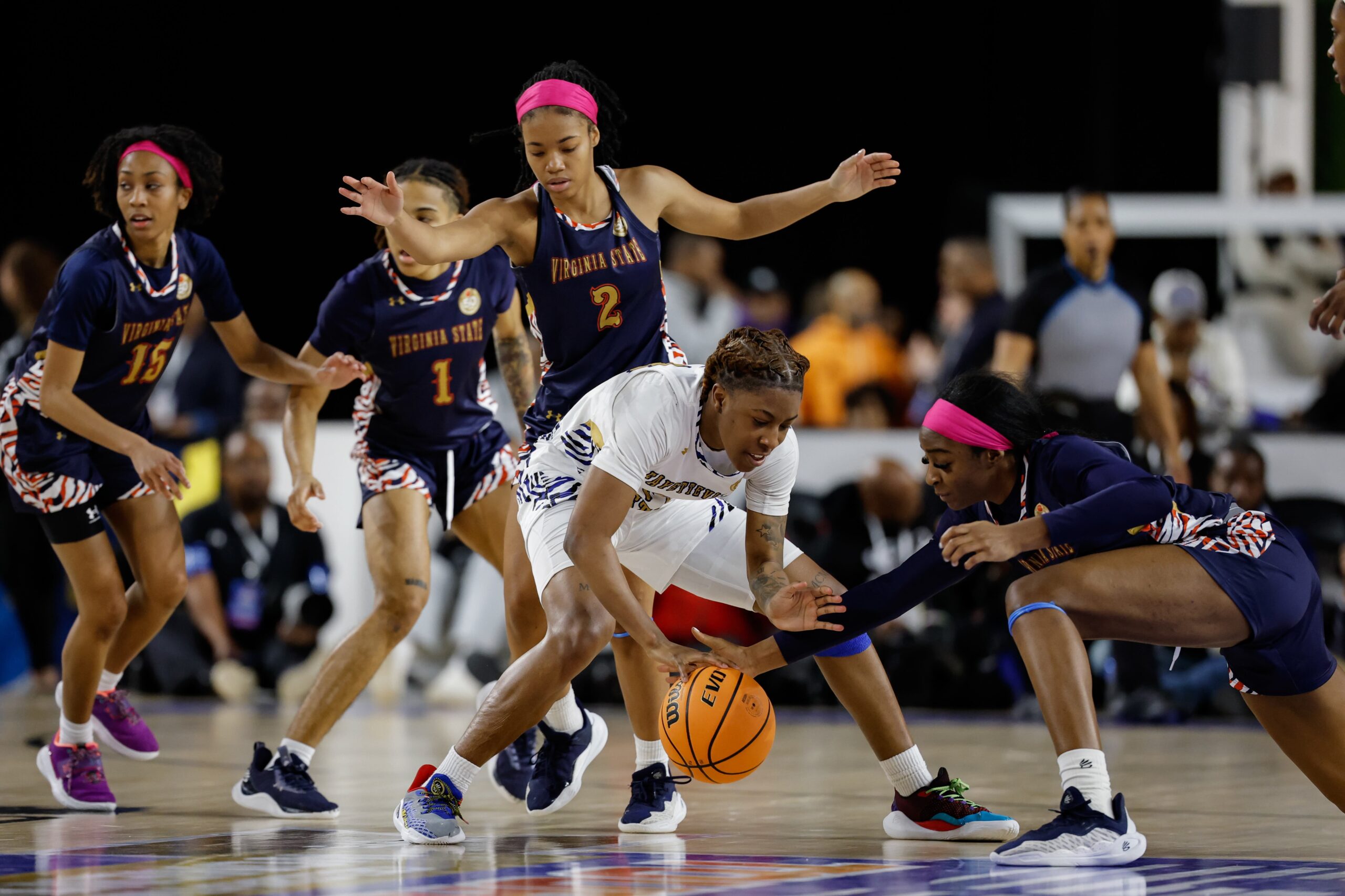 Players from two basketball teams compete for the ball on a court. One player in white reaches to secure the ball as players in blue and orange attempt to intercept.