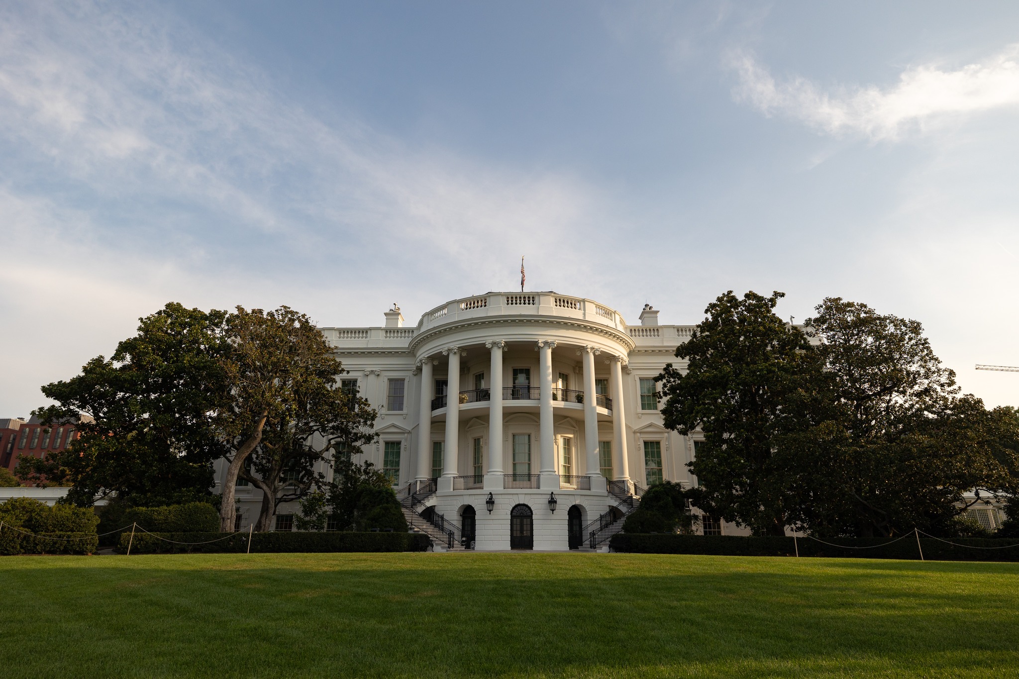 The White House's south facade with columns, viewed from the lawn, surrounded by trees under a partly cloudy sky.