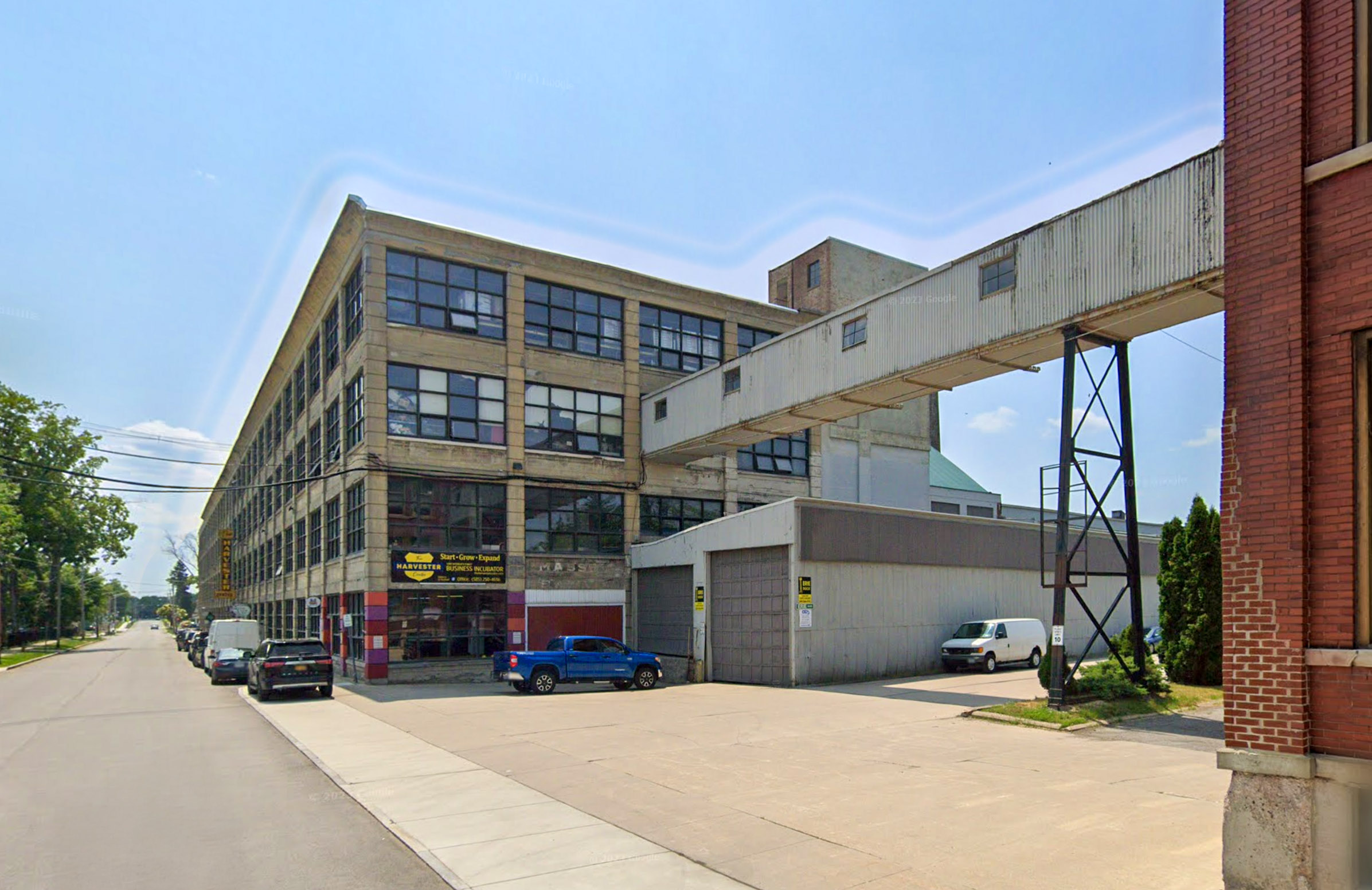 A multi-story industrial building with large windows and a connecting walkway on a street with parked cars and clear skies.