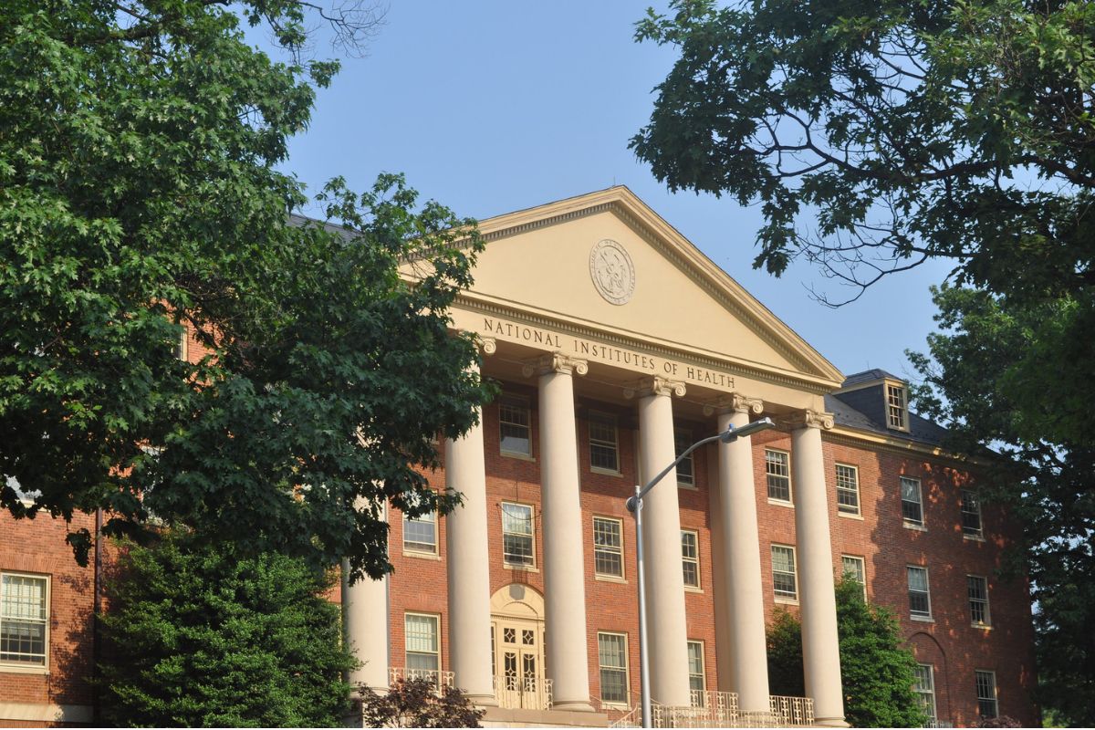A brick building with tall columns and "National Institutes of Health" inscribed on the facade, partially obscured by trees, under a clear blue sky.