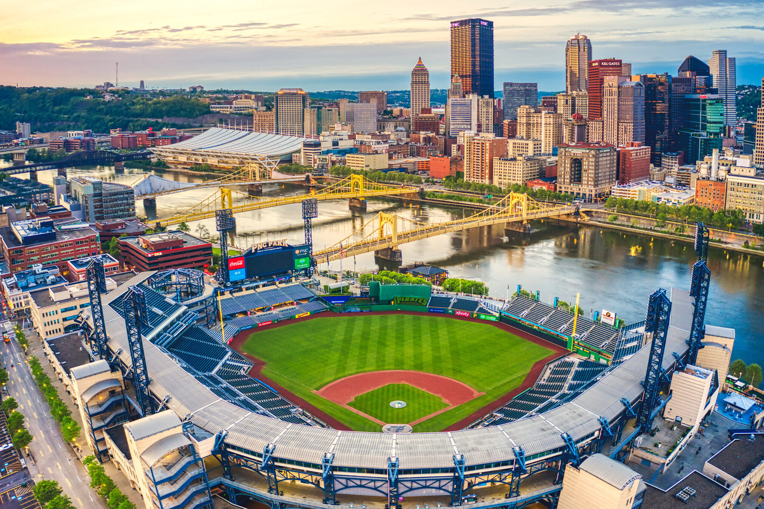 Aerial view of a baseball stadium with a city skyline and river in the background, showing greenfield and seating areas, surrounded by urban streets and buildings.