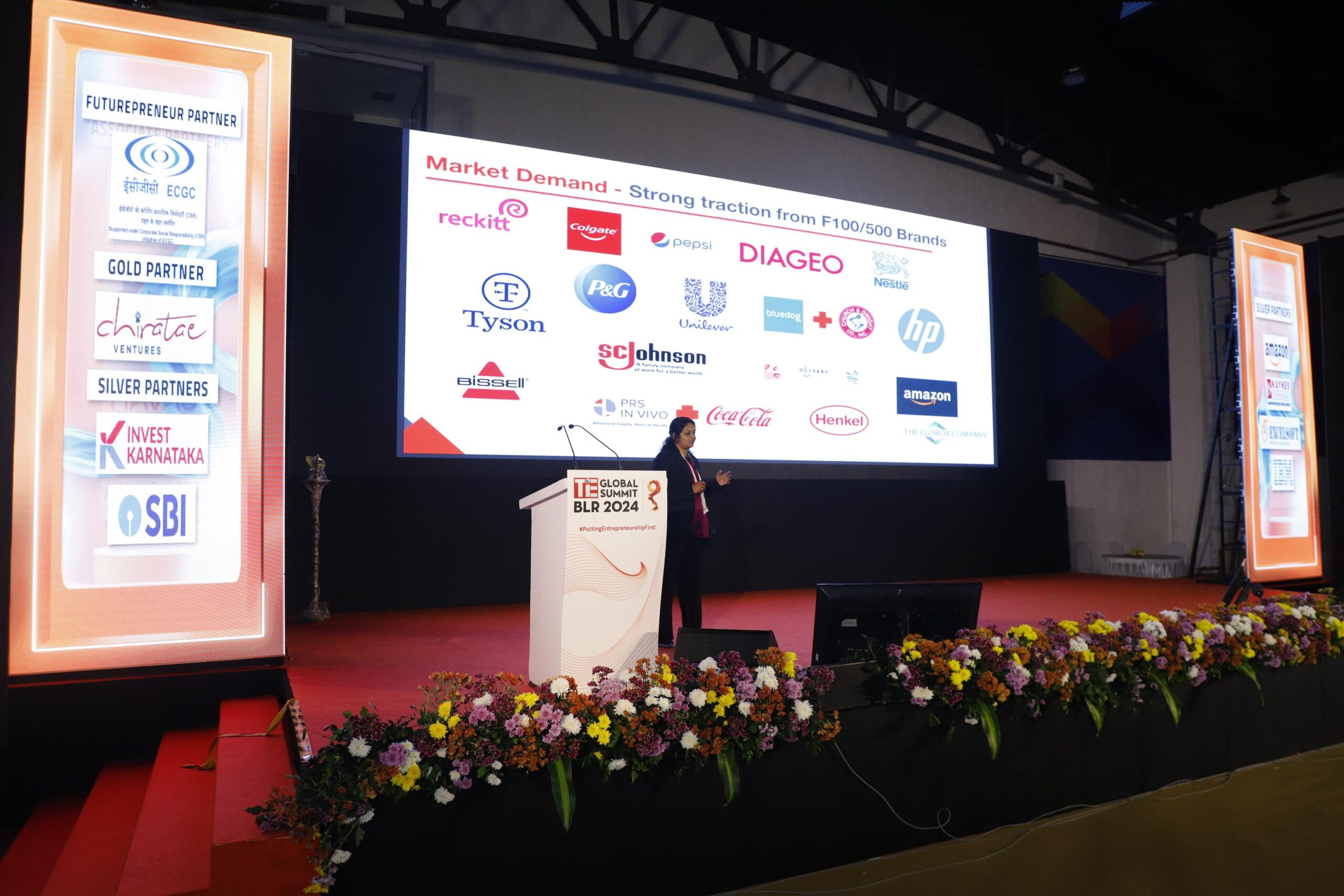 A woman speaks at a podium on a stage with a large screen displaying logos of various brands like Coca-Cola, HPand Amazon. The stage is decorated with flowers.