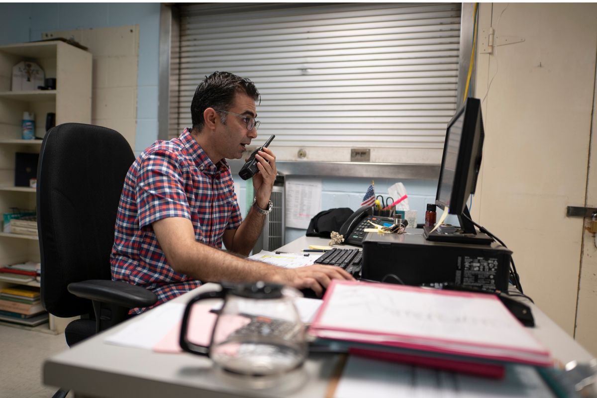 A man in a plaid shirt is sitting at a desk, speaking into a handheld radio while looking at a computer screen, surrounded by office supplies in a room with shelves and a metal shutter.