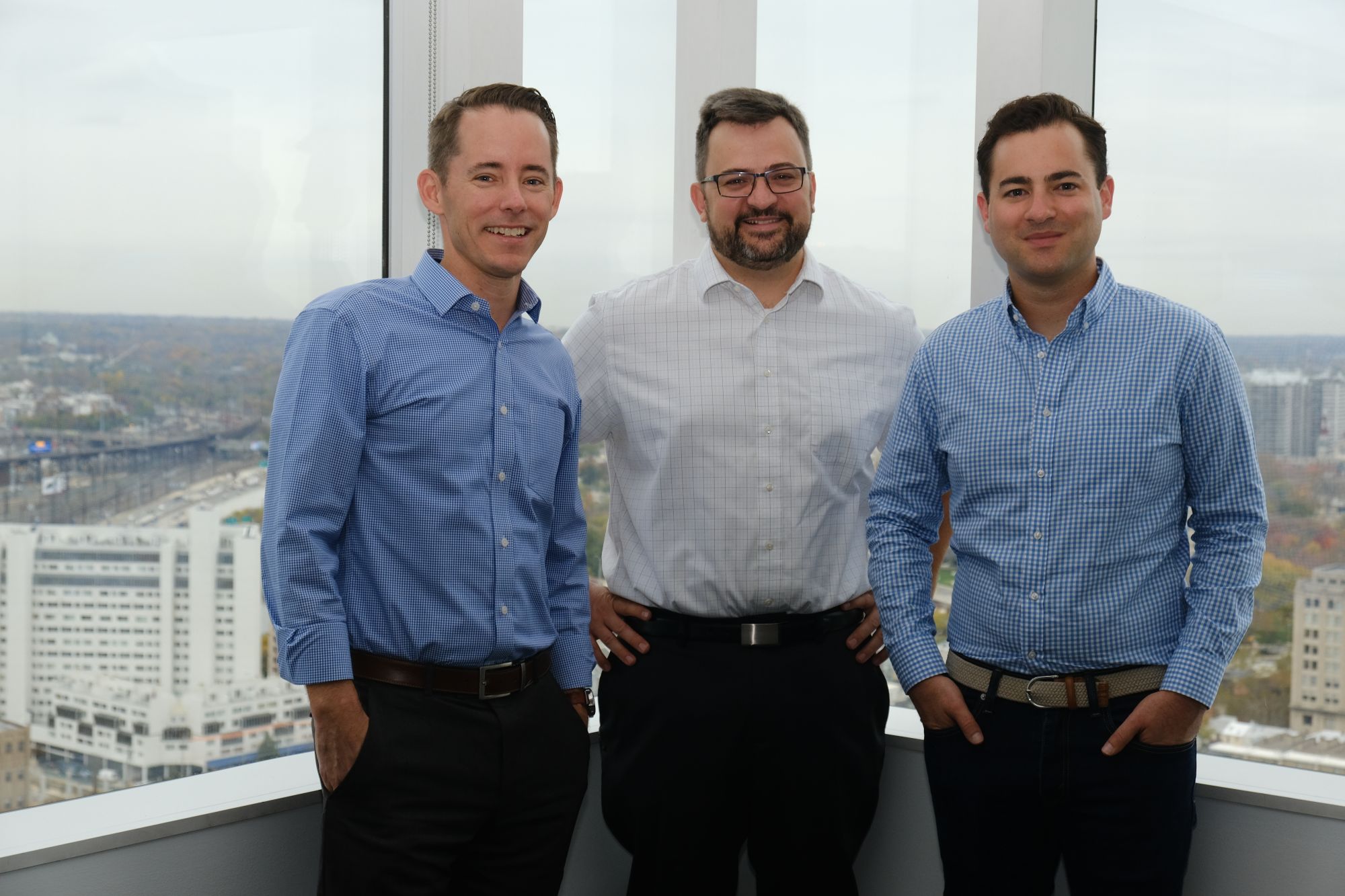 Three men in button-up shirts stand in an office with a cityscape view in the background.