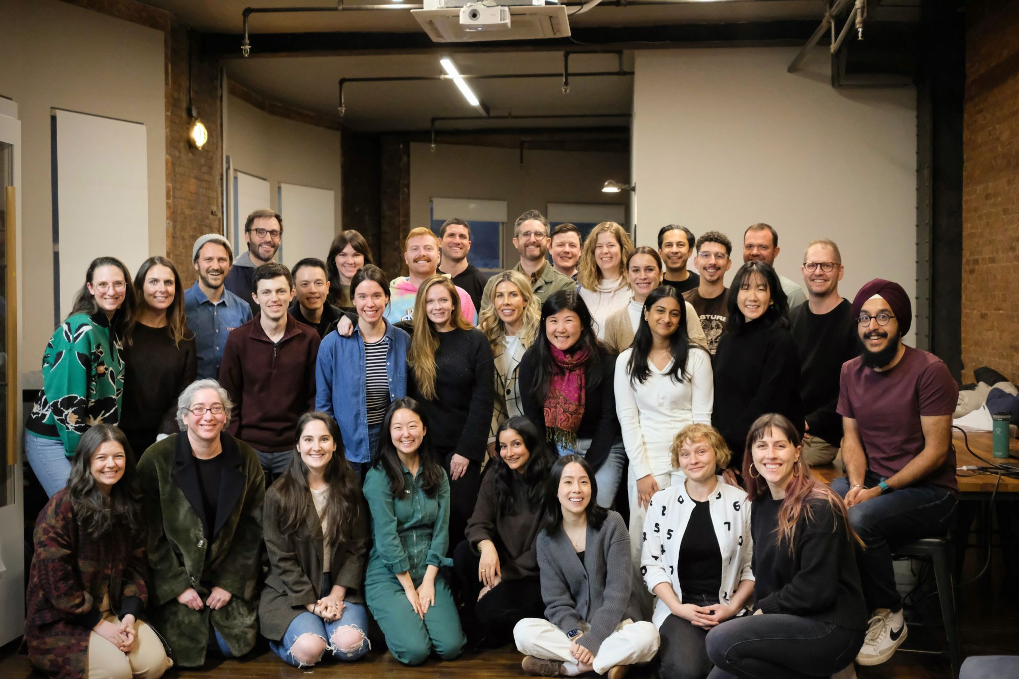 A group of about 30 people pose together indoors, smiling at the camera in a casual office setting with exposed brick walls.