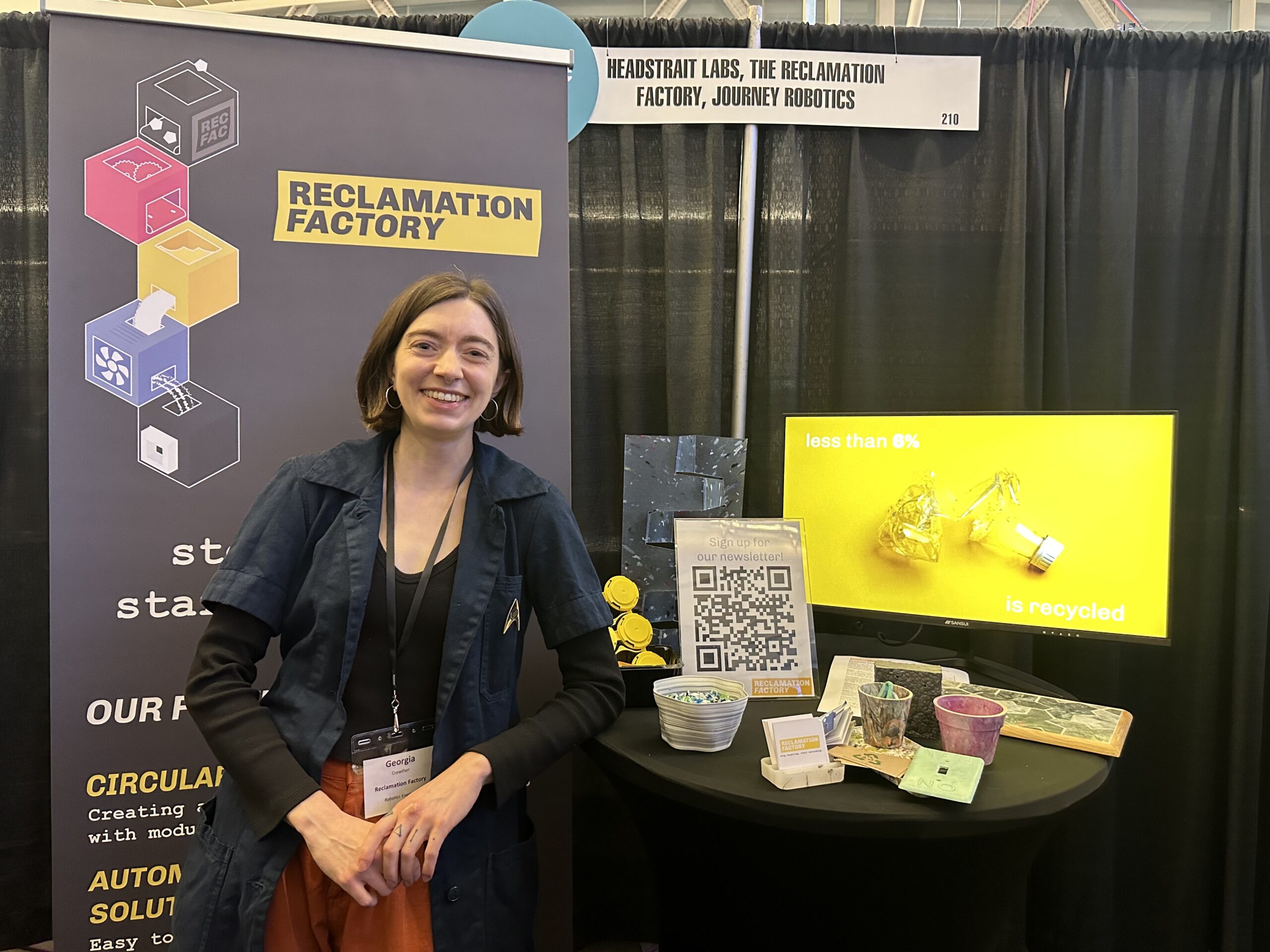 A person stands in front of a booth for Reclamation Factory, featuring a sign, QR code, samples and a display about recycling at an event.