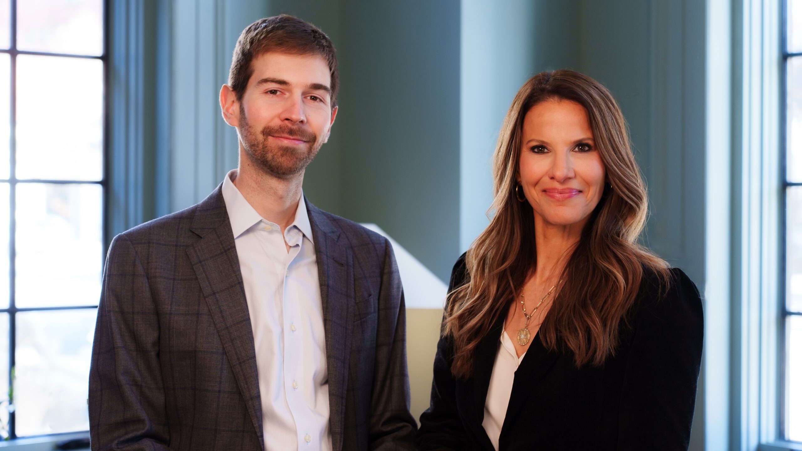 A man and a woman in business attire stand side by side, smiling confidently at the camera.
