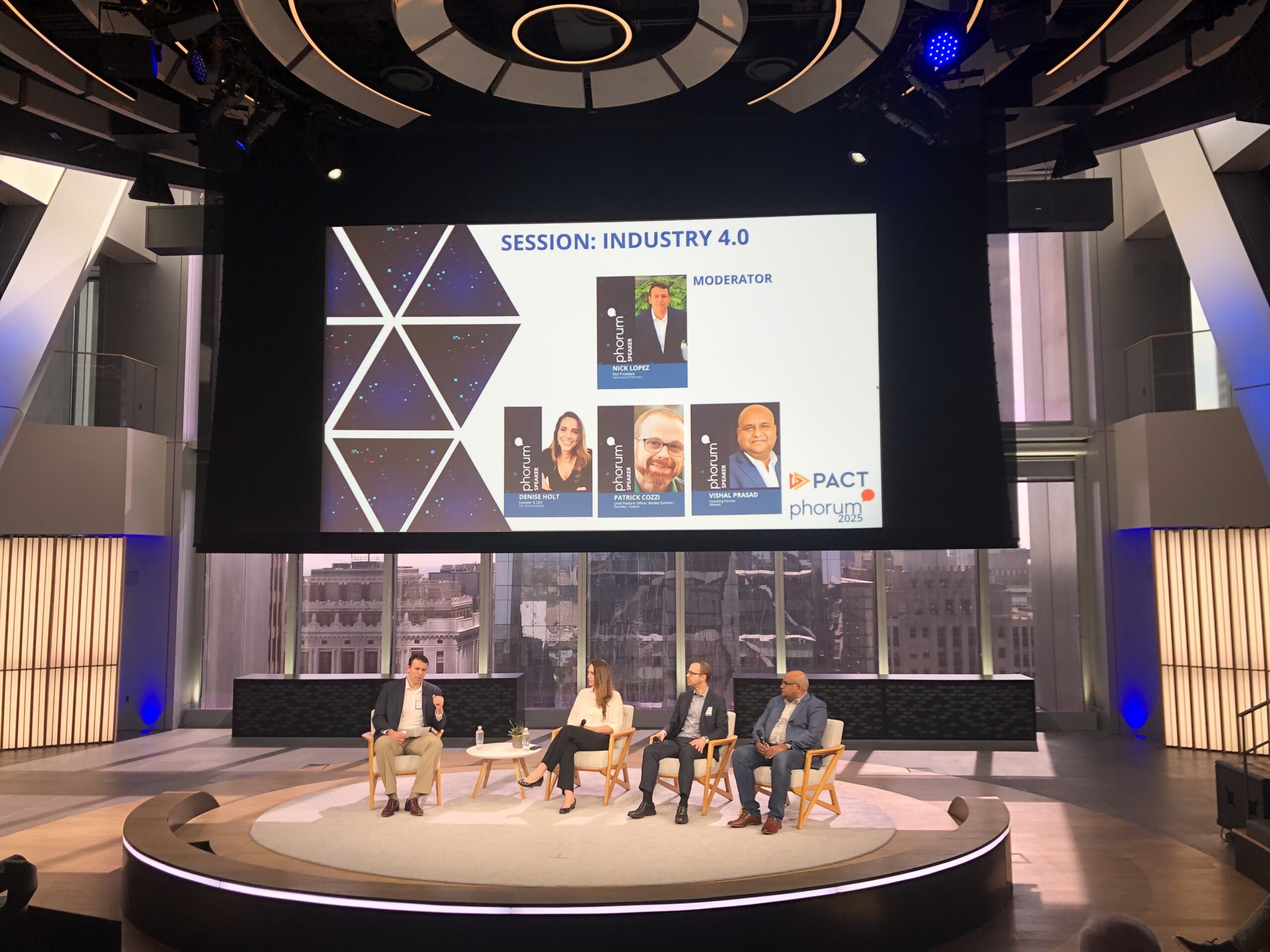 Four people sit on stage panel under a large screen displaying "Session: Industry 4.0" with moderator and speaker names and photos at a professional event in a modern venue.