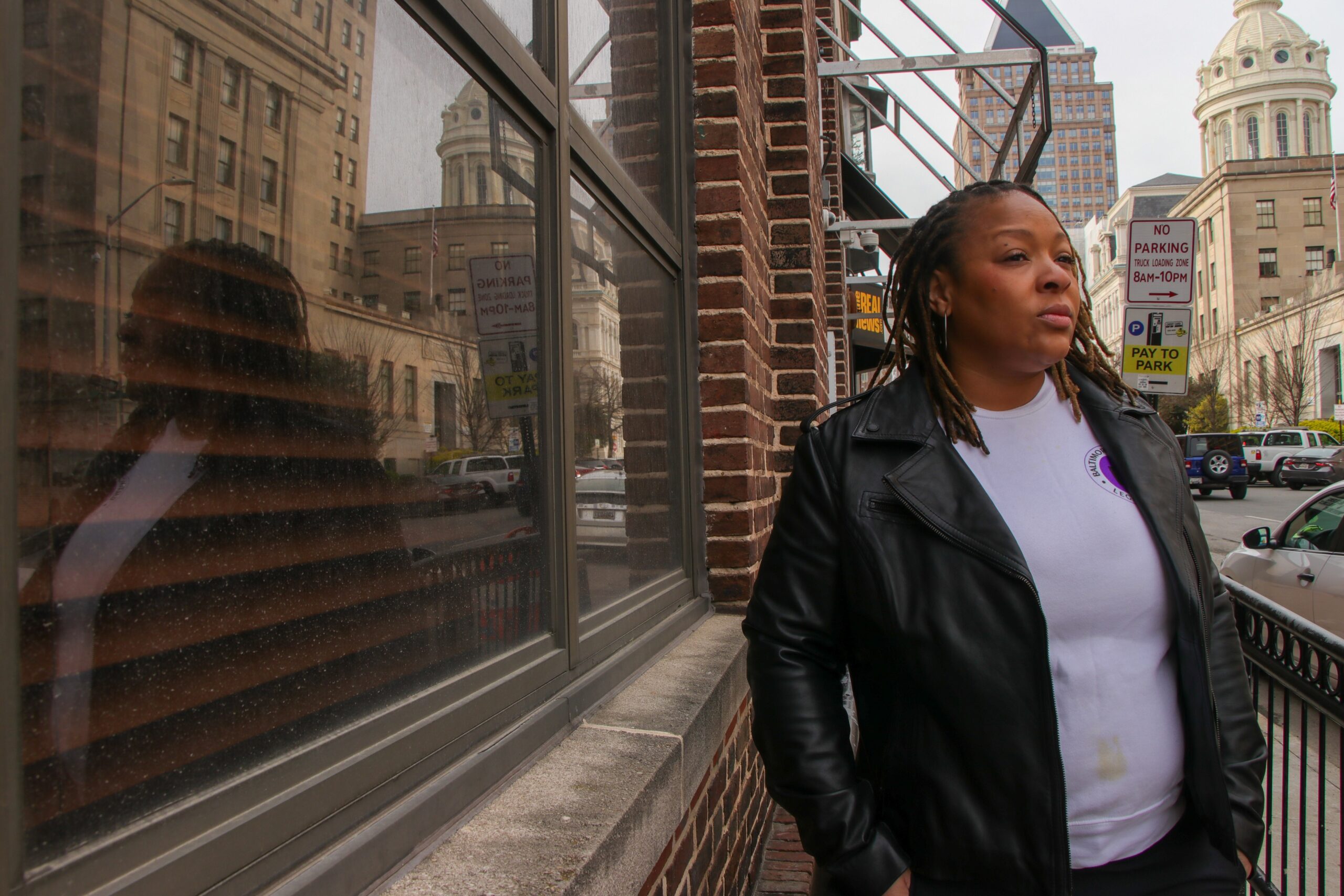 A woman in a black leather jacket stands on a city sidewalk next to a brick building, with her reflection visible in the window. City buildings and street signs are in the background.