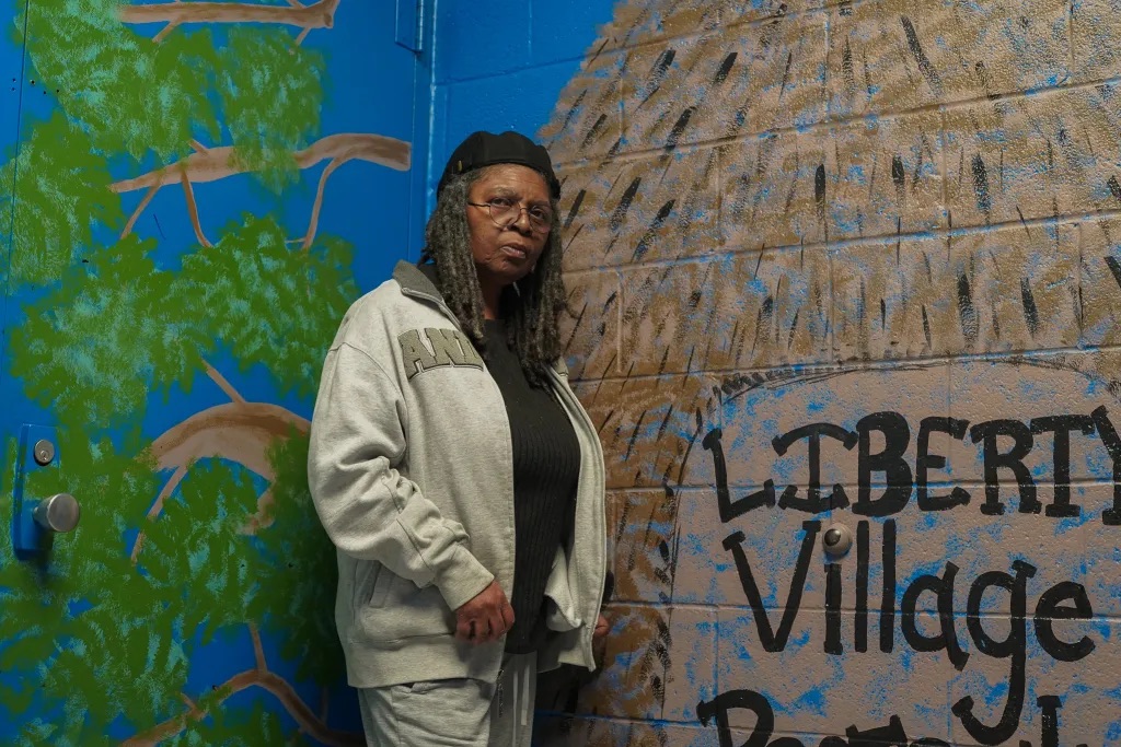 A woman wearing a gray tracksuit and black hat stands in front of a painted mural that says "LIBERTY Village" on a brick wall.