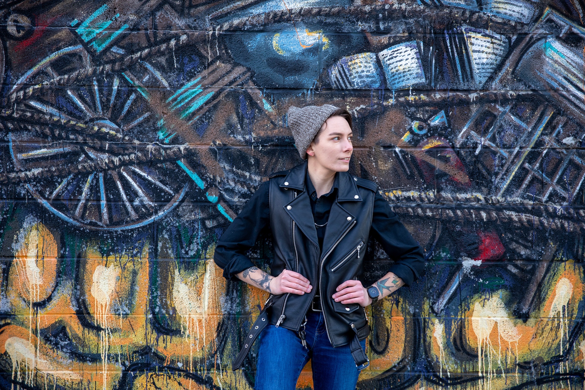 A person in a gray beanie and black leather vest stands in front of a colorful mural featuring wheels, books, and abstract patterns.