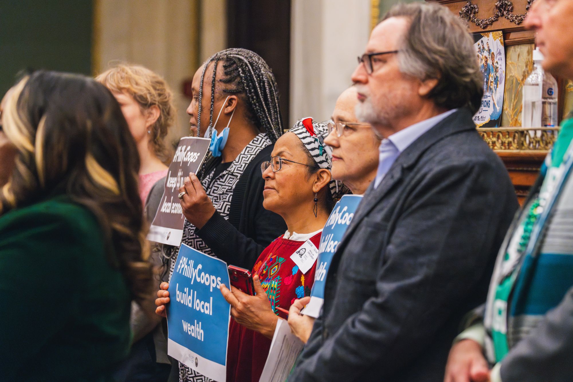 A group of people stand in a room holding signs that say "Coops build local wealth." They are listening attentively.