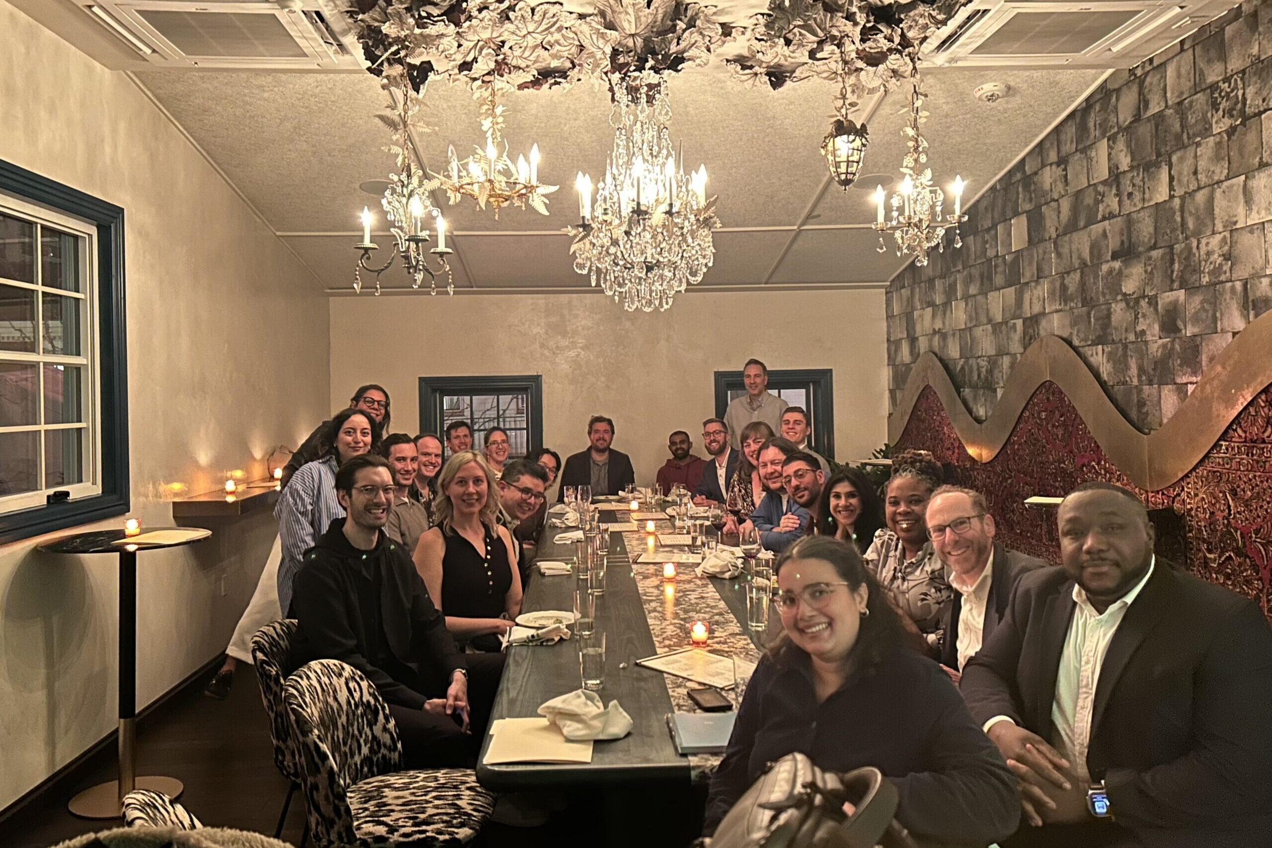 A group of people sit around a long dining table in a dimly lit restaurant with a chandelier above them.